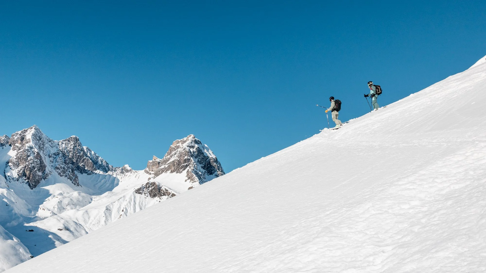 Two skiers ascending a snowy mountain slope with a backdrop of rugged, snow-covered mountains under a clear blue sky.