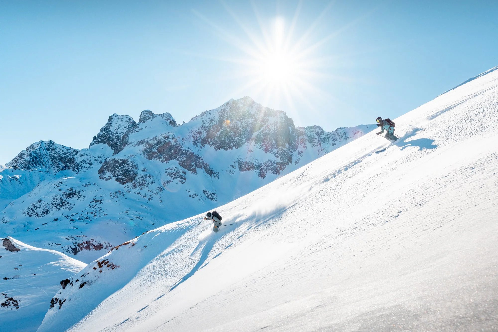 Two skiers descending a snowy mountain slope with mountain peaks in the background and the sun shining brightly overhead.