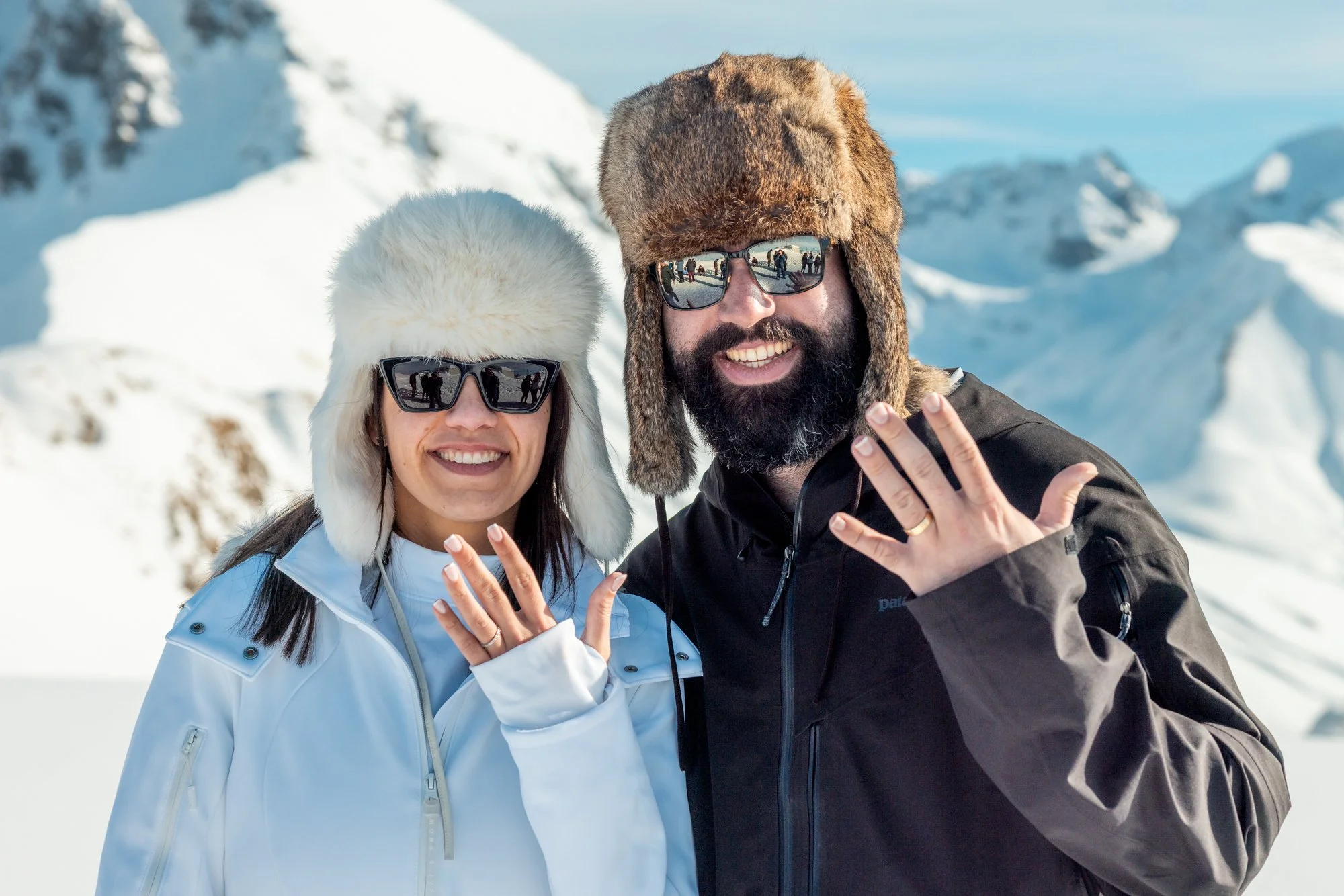 A smiling couple in winter clothing and sunglasses showing off their rings, standing in snowy mountains.