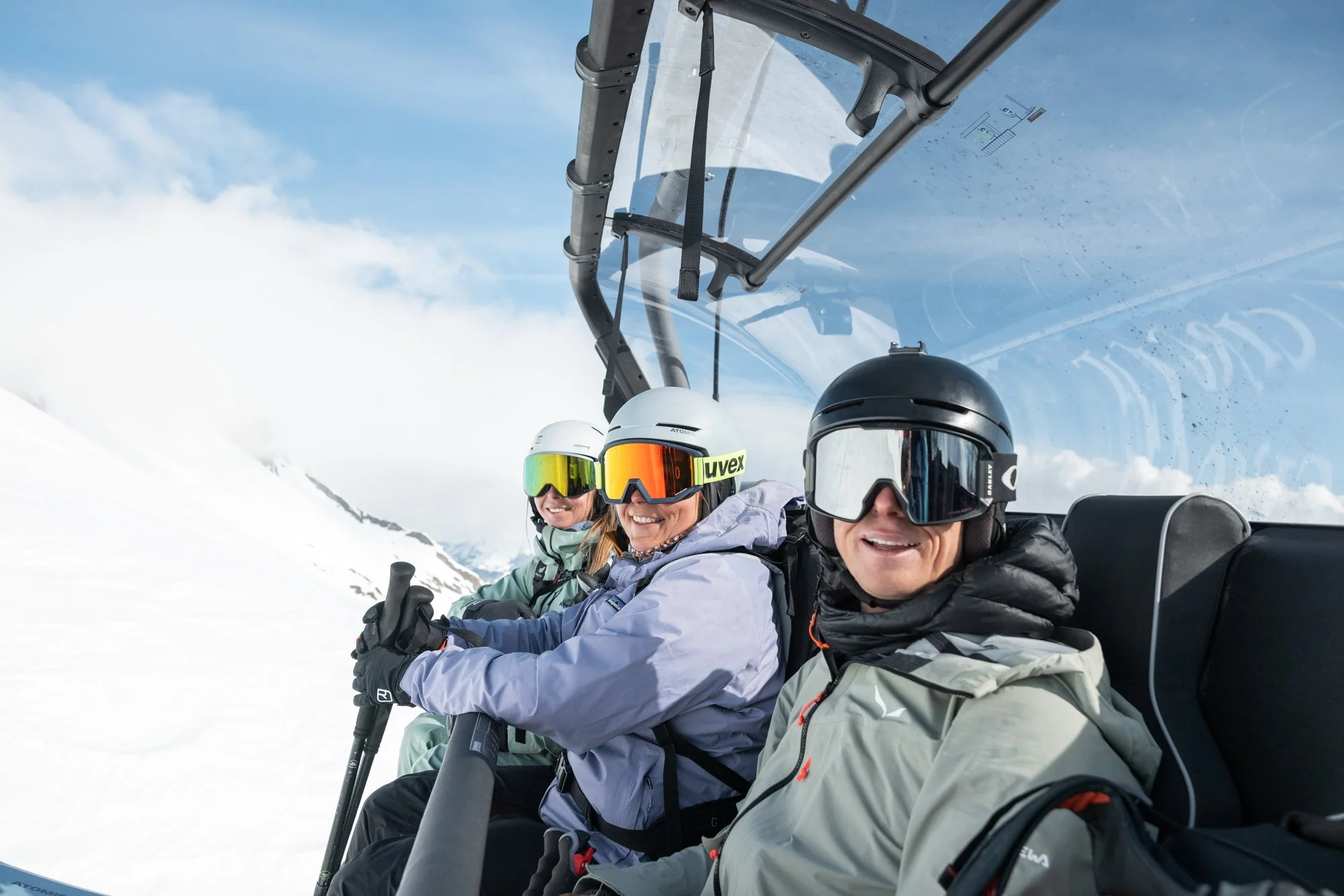 Three skiers in ski helmets and goggles sitting in a ski lift chair above snow-covered mountains and blue sky, smiling.