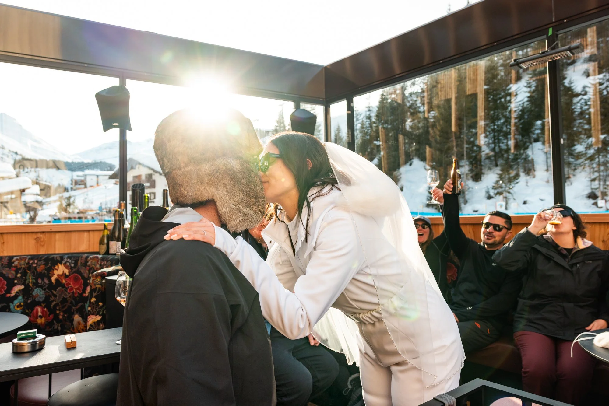 A woman in white jacket is kissing a man with sunglasses and a fur hat in a mountain restaurant during sunny day, with friends in the background celebrating.
