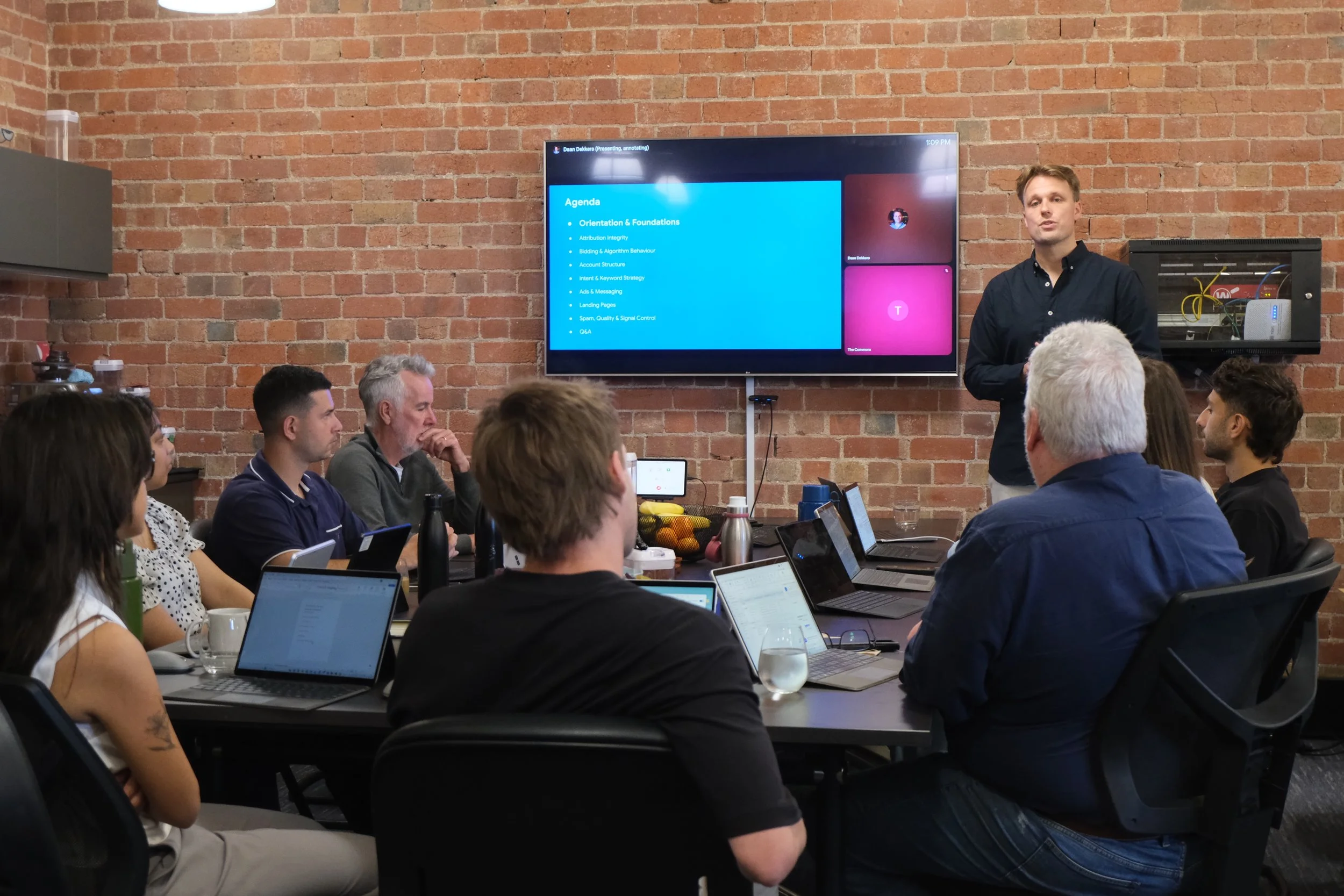 Overhead view of a person working at a white desk with a laptop, smartphone, coffee mug with latte art, remote control, phone on charging dock, notepad, and pens.