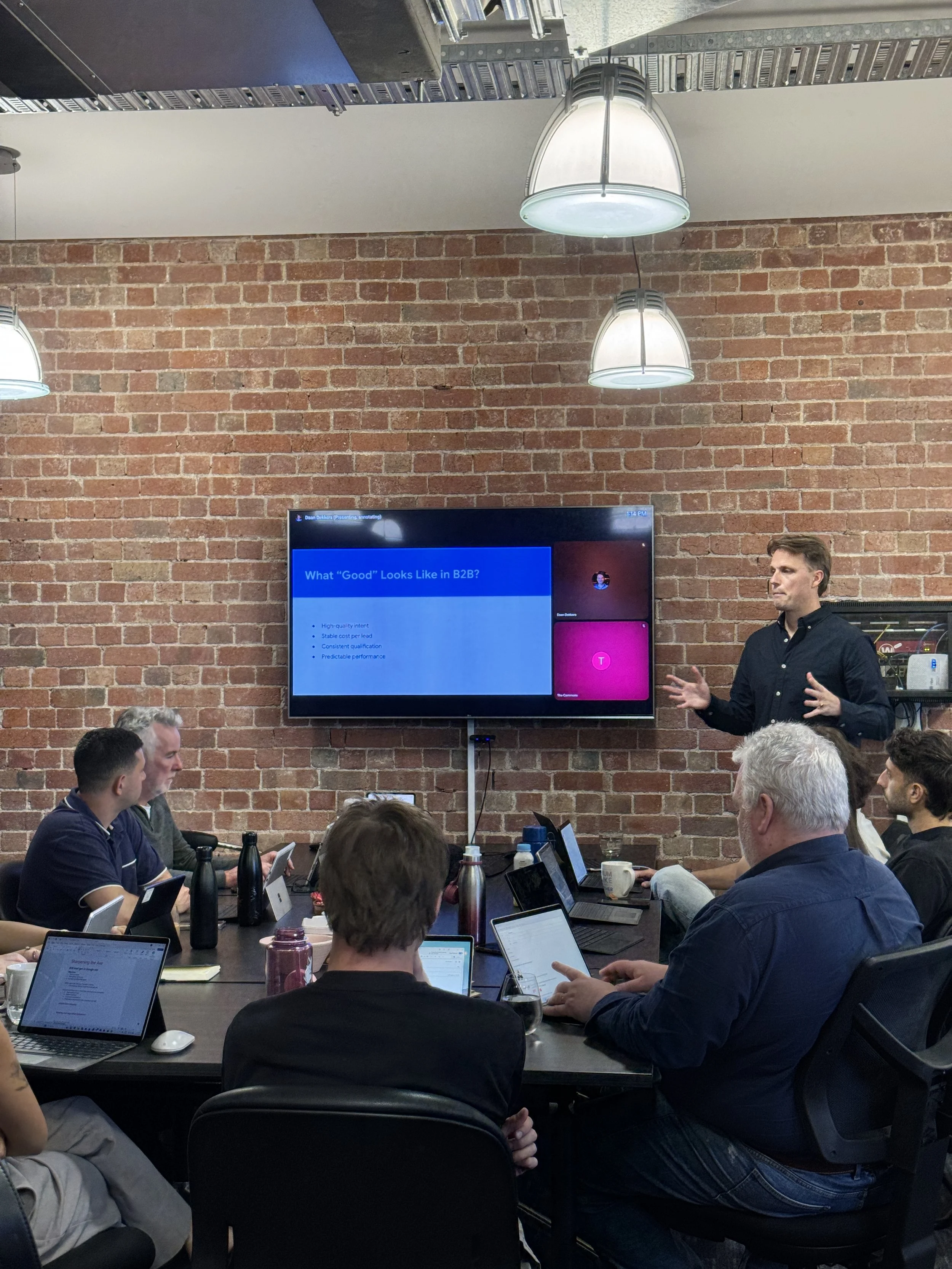 A group of five young adults sitting at a wooden table in a rustic room with large windows, working on laptops and wearing headphones.