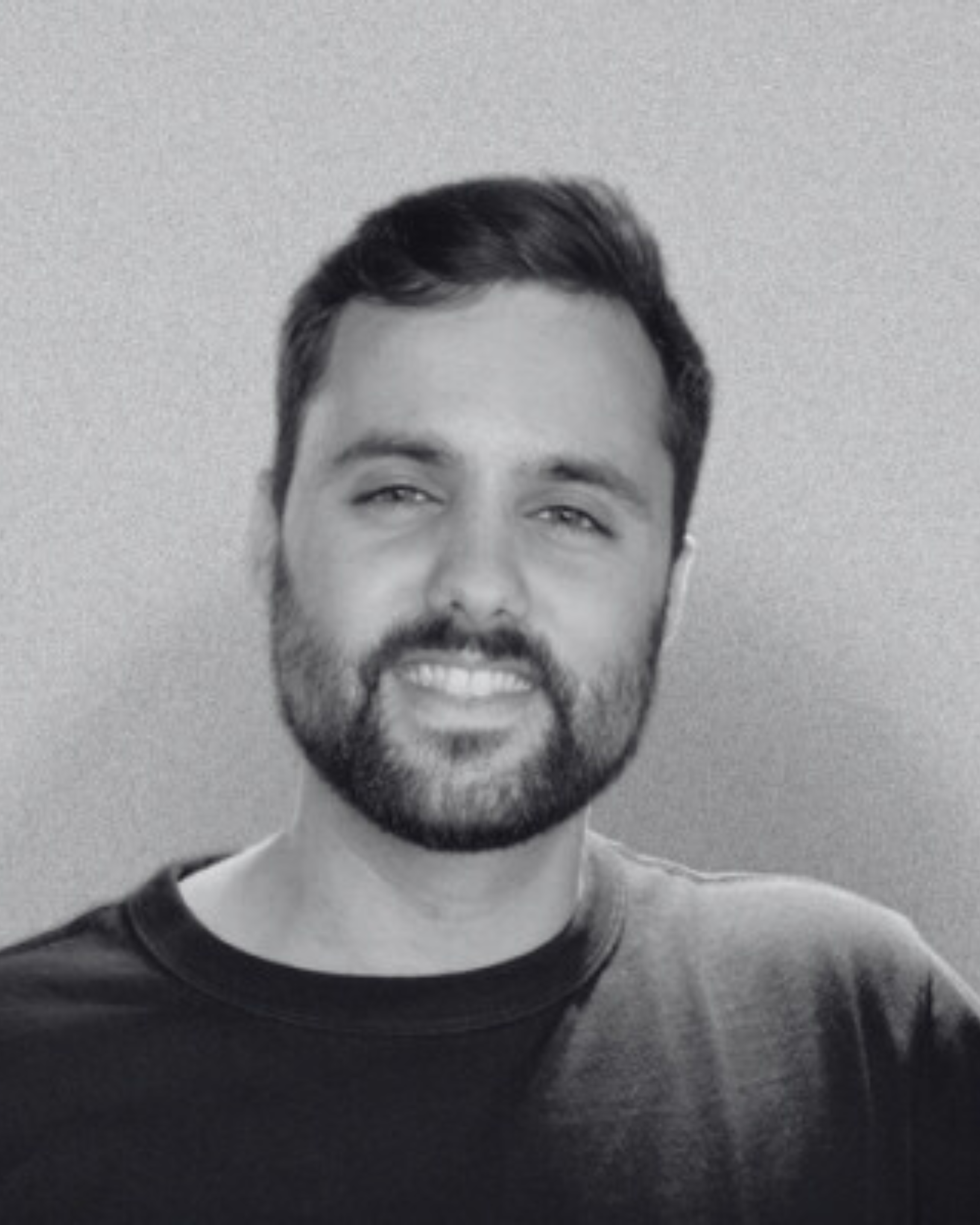 Black and white photograph of a man with dark hair, facial hair, smiling, wearing a dark T-shirt, standing against a plain background.