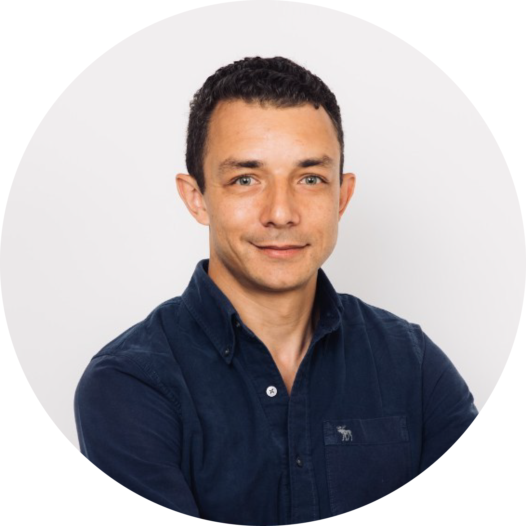 Portrait of a young man with short dark hair and blue eyes wearing a dark blue collared shirt, smiling against a plain white background.