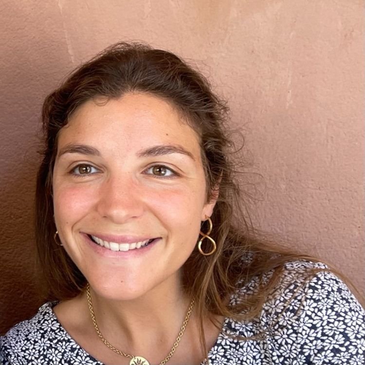 A young woman with brown hair, wearing hoop earrings and a necklace, smiling against a tan-colored wall.