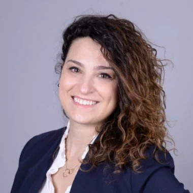 Portrait of a woman with curly brown hair, smiling, wearing a dark blazer and a white shirt.
