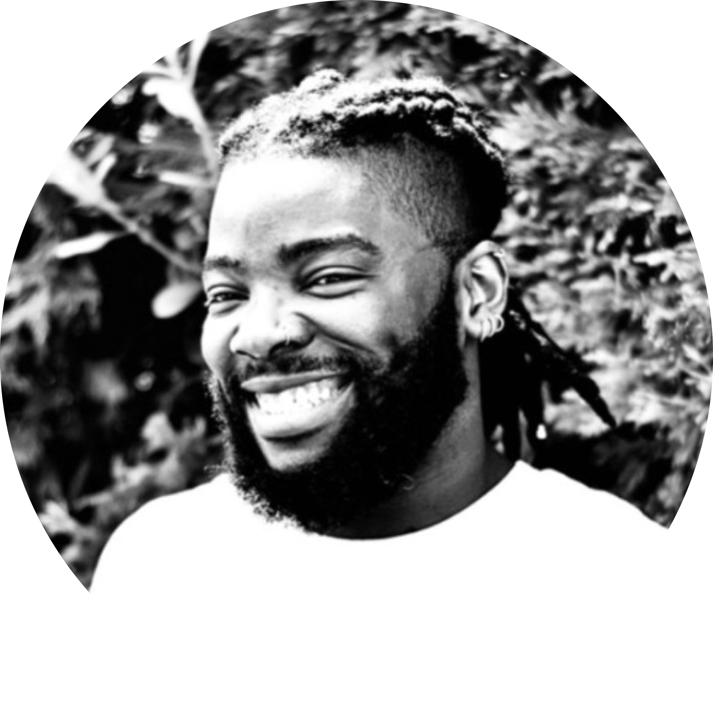 Black and white photo of a smiling man with a beard and short dreadlocks, outdoors with foliage in the background.