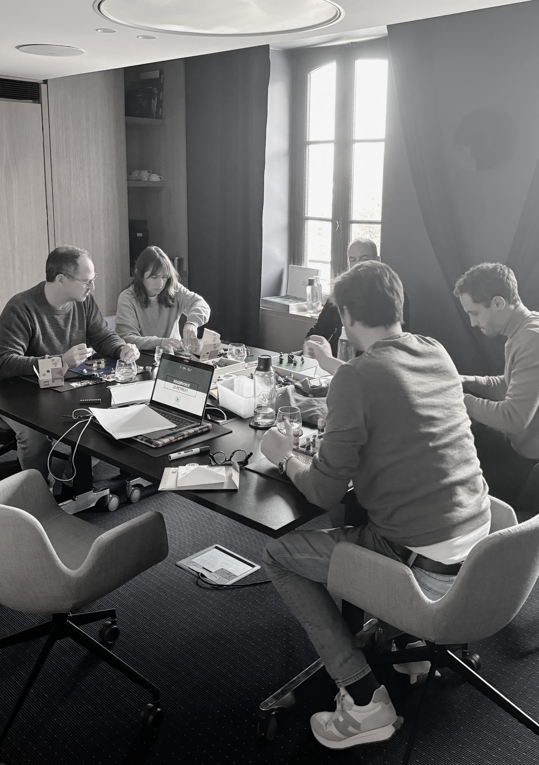 Five people playing chess at a table in a bright room with large windows, some on laptops, with scattered papers and glasses on the table.