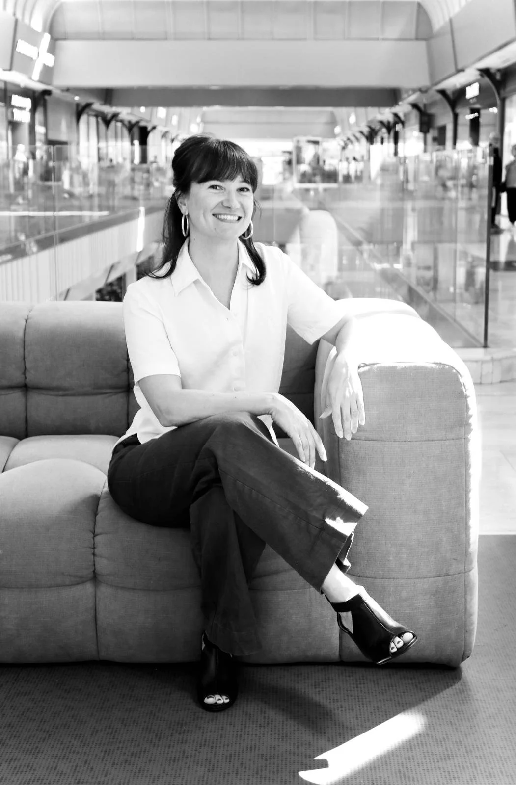 A woman sitting on a sofa in an airport terminal, smiling at the camera. She is wearing a white blouse, dark pants, and open-toe heels.