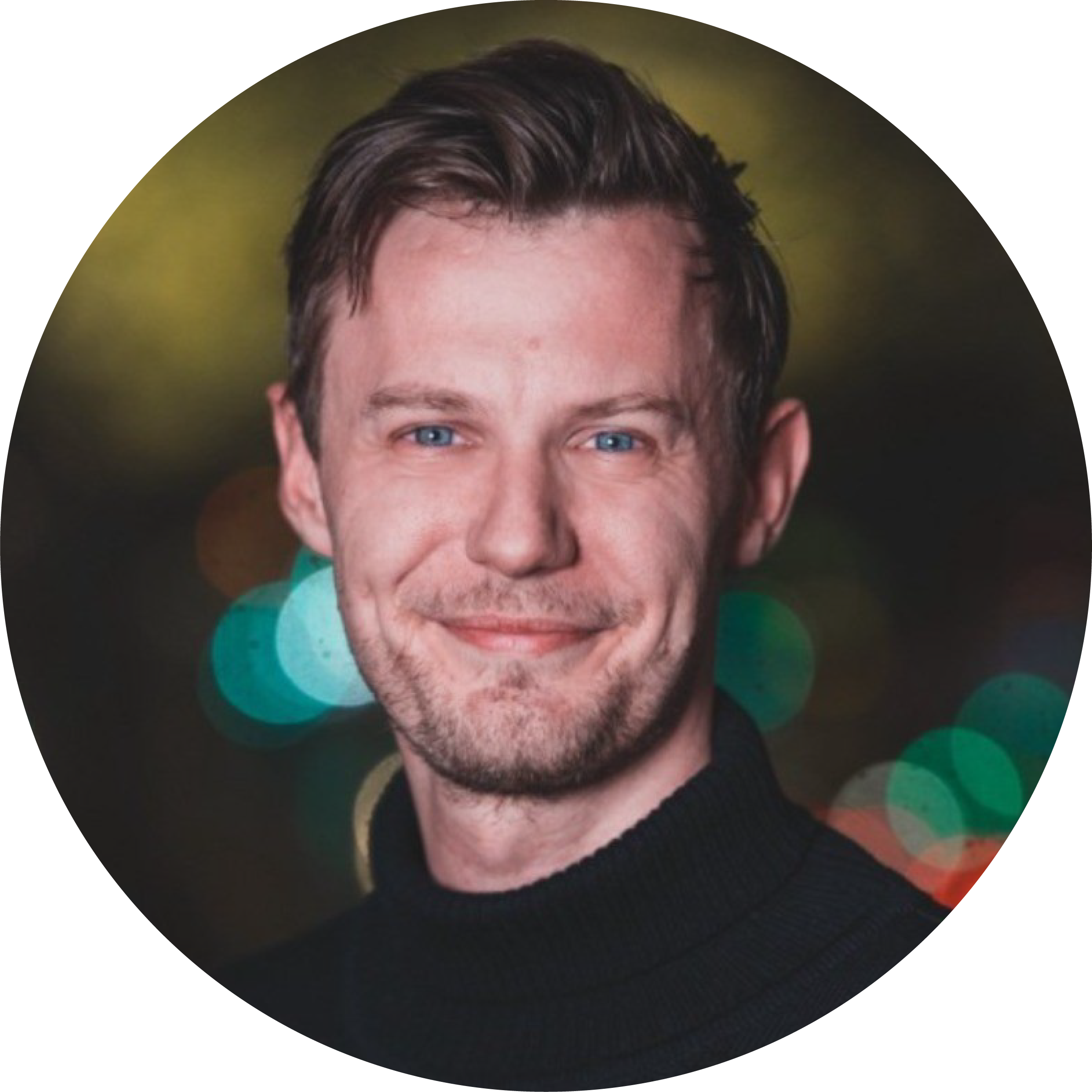 Portrait of a man with short, dark hair and blue eyes, smiling, wearing a black turtleneck, with blurred colorful city lights in the background.