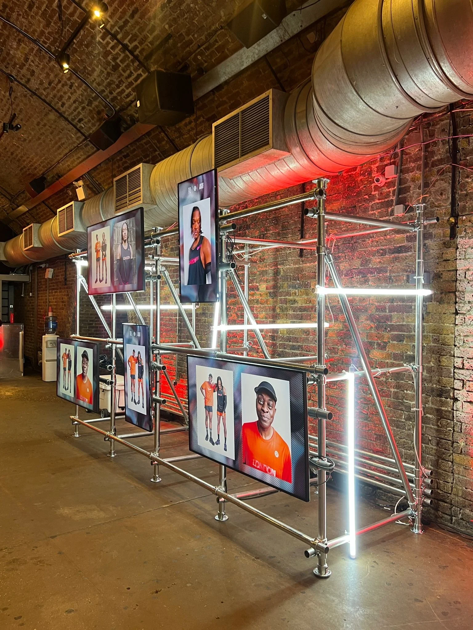 Interior of a cafe or coffee shop with posters of women in athletic wear on a brick wall, barista working behind the counter with coffee equipment, menu board listing teas and breakfast items, and warm lighting.