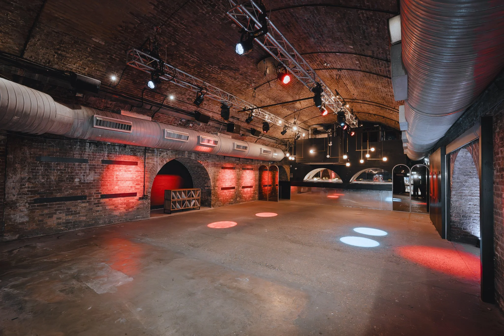 Empty dance floor with colored spotlights on the floor, exposed brick walls, and professional lighting equipment overhead in a venue or club.
