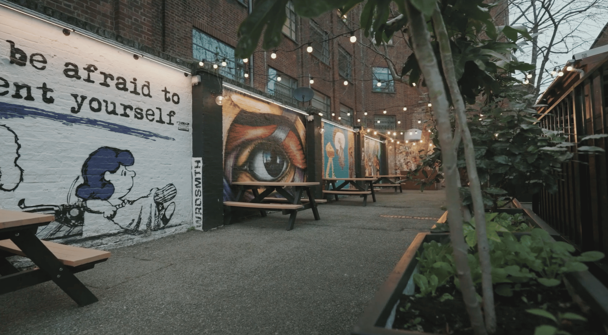 An outdoor patio area with string lights, wooden benches, and colorful murals on a brick wall, featuring an eye and cartoon characters.