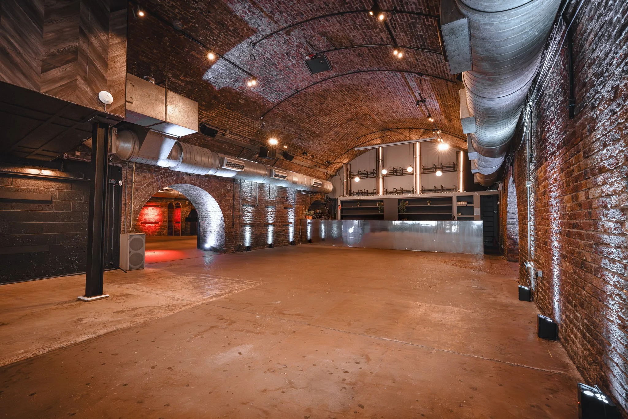 An empty, industrial-style indoor space with exposed brick walls, a vaulted brick ceiling, and visible ductwork. The room is lit with multiple ceiling spotlights and has a polished concrete floor. There is a bar or counter area in the background, and the space appears to be designed for events or gatherings.
