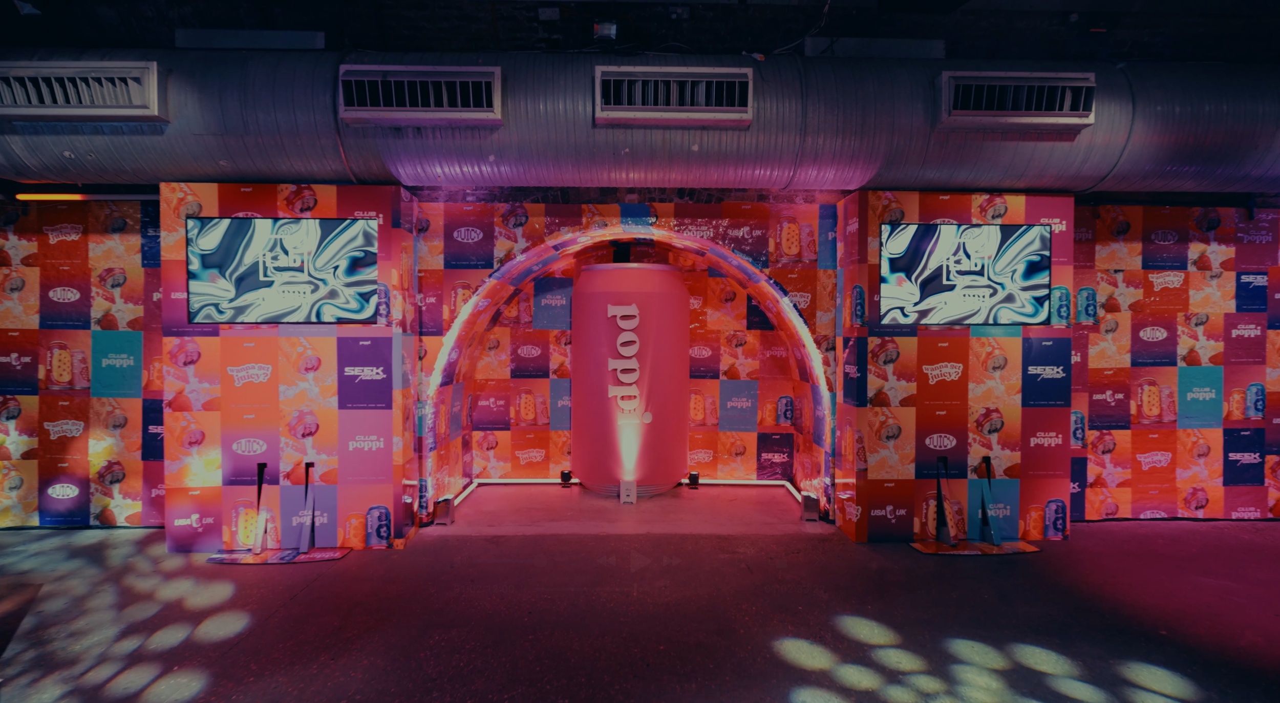 A colorful event setup with walls decorated in bright pink, purple, and orange patterns featuring product advertisements. There are two large screens with swirling abstract visuals, and a large pink pillar in the center with the word 'daddo' written vertically. Overhead metallic ceiling vents are visible, and the floor is illuminated with spotlights creating circular light patterns.