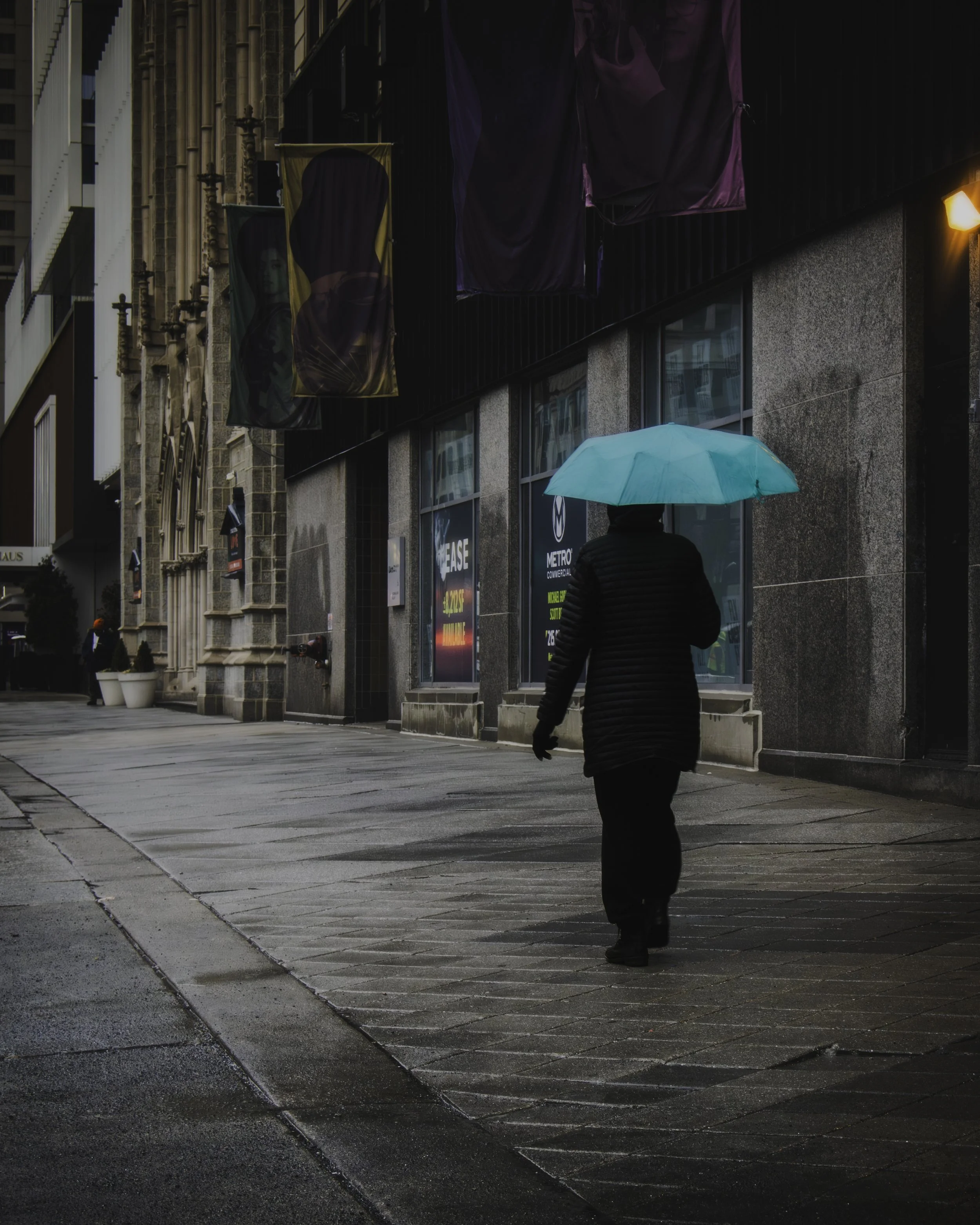 Quiet Block; Along the Facade
Person carrying a pale blue umbrella walks alone along a rain-darkened sidewalk lined with banners and stone facades.