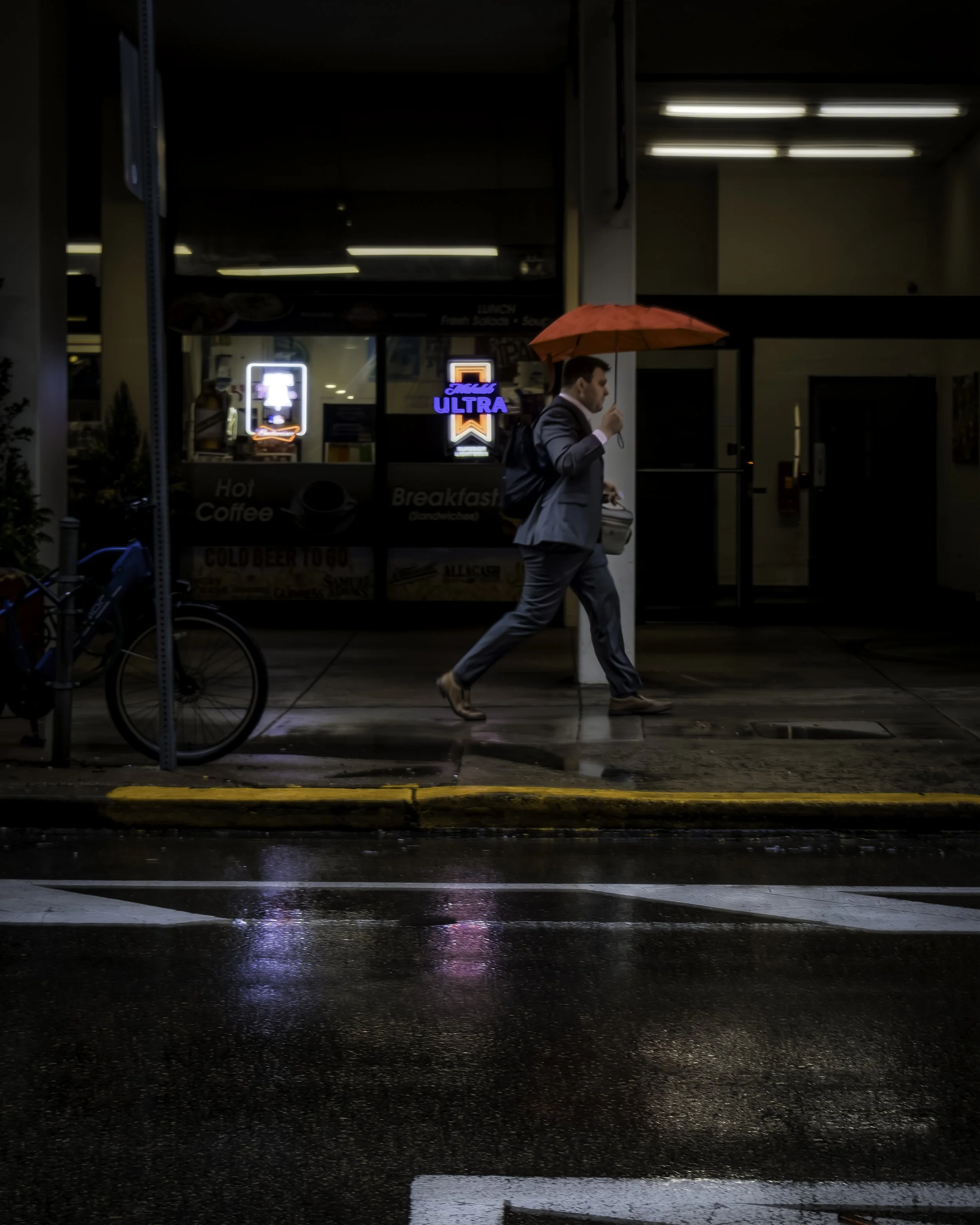Red Umbrella Morning
Person walking with an orange umbrella on a rainy city street, framed by storefront lights and reflections on wet pavement.