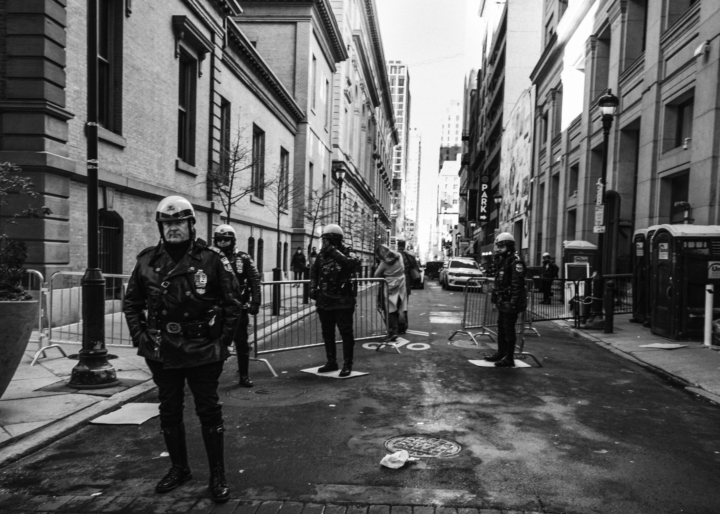 Street of Authority
Black and white street scene on New Year’s Day with several police officers standing behind metal barricades on a wet roadway.