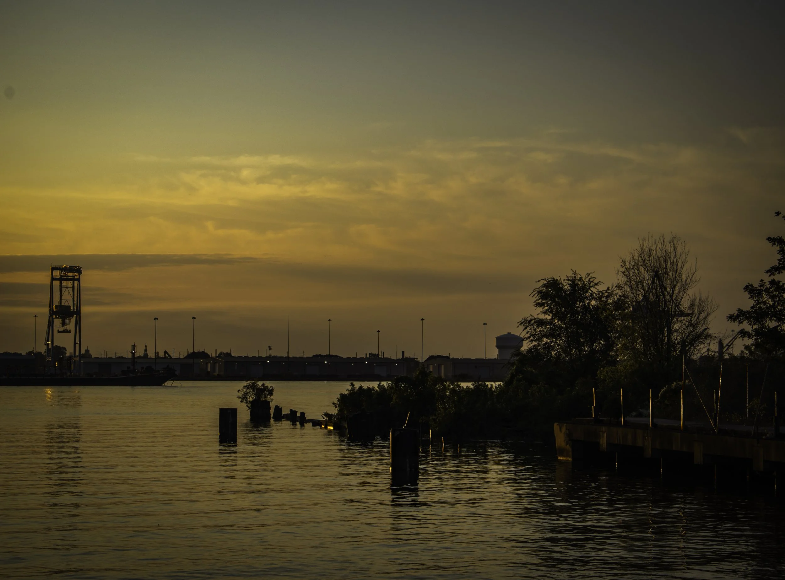 Golden Edge of Morning
Calm river at sunrise with industrial silhouettes, dock posts, and trees along the right edge beneath a wide golden sky.