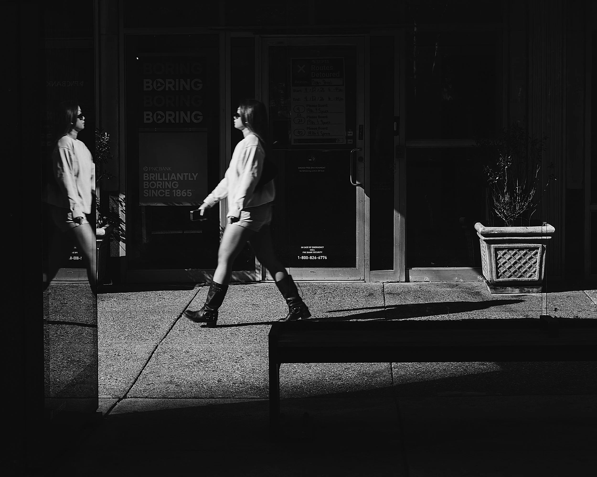 Brilliantly Not Boring
Black-and-white street photo of a woman walking past a bus stop bench and glass storefront doors, with her reflection visible in the glass and strong shadows across the sidewalk.