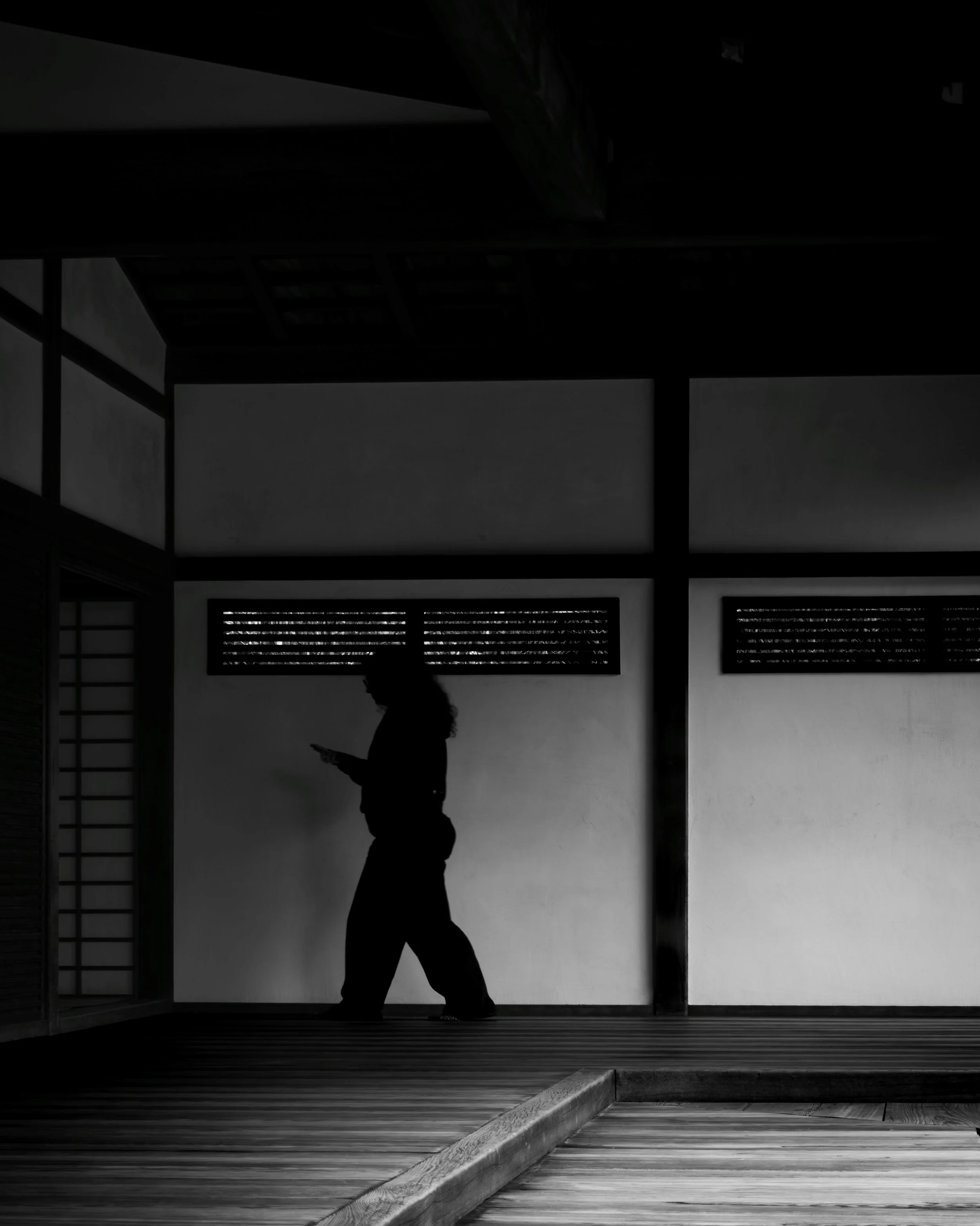 Between Walls and Thought
Black-and-white photograph of a silhouetted woman walking through a Japanese-style wooden corridor while looking at her phone, with bright wall panels behind her.