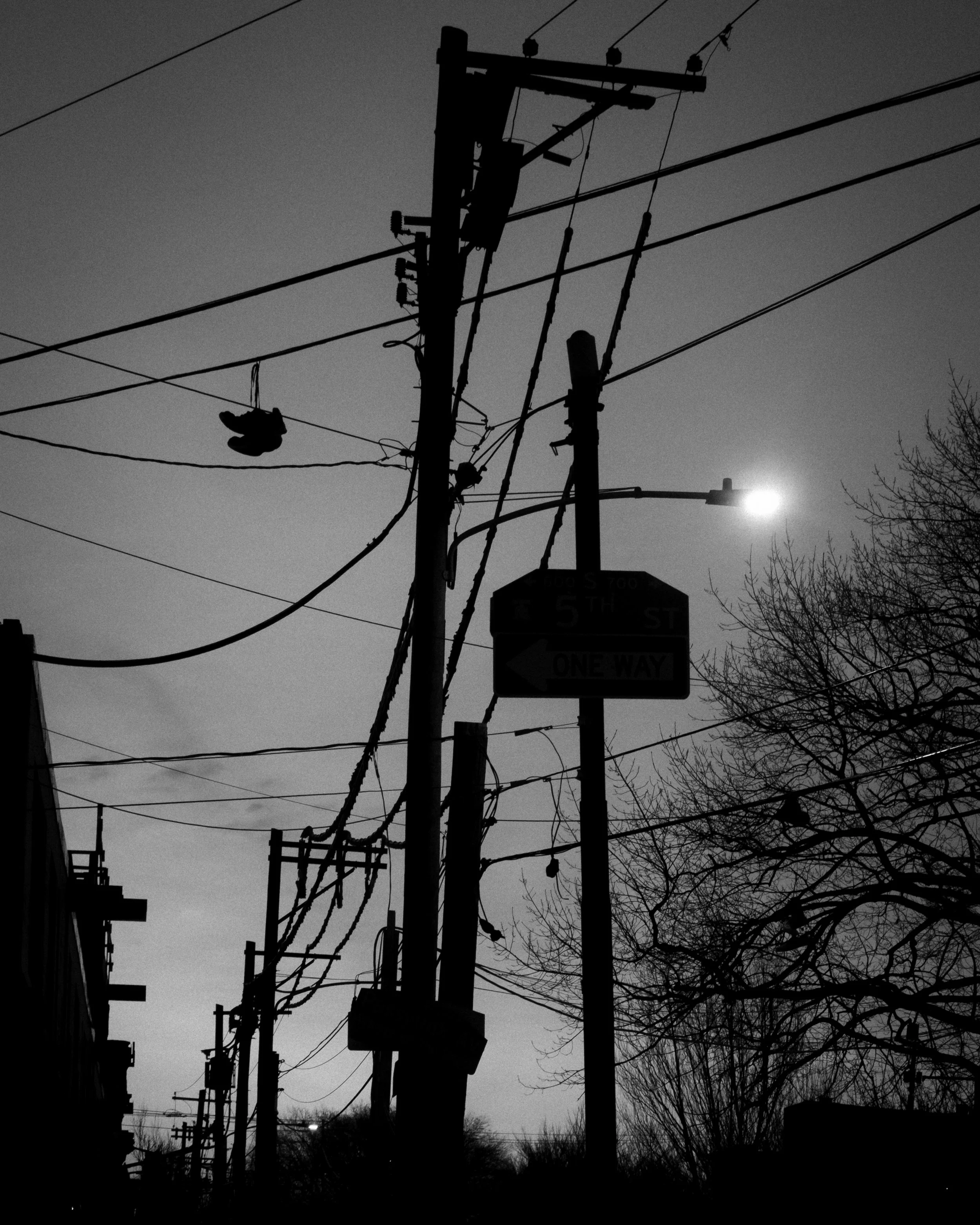 Hanging Above the Block
Black and white urban scene of utility poles, tangled power lines, hanging sneakers, and a one-way sign.