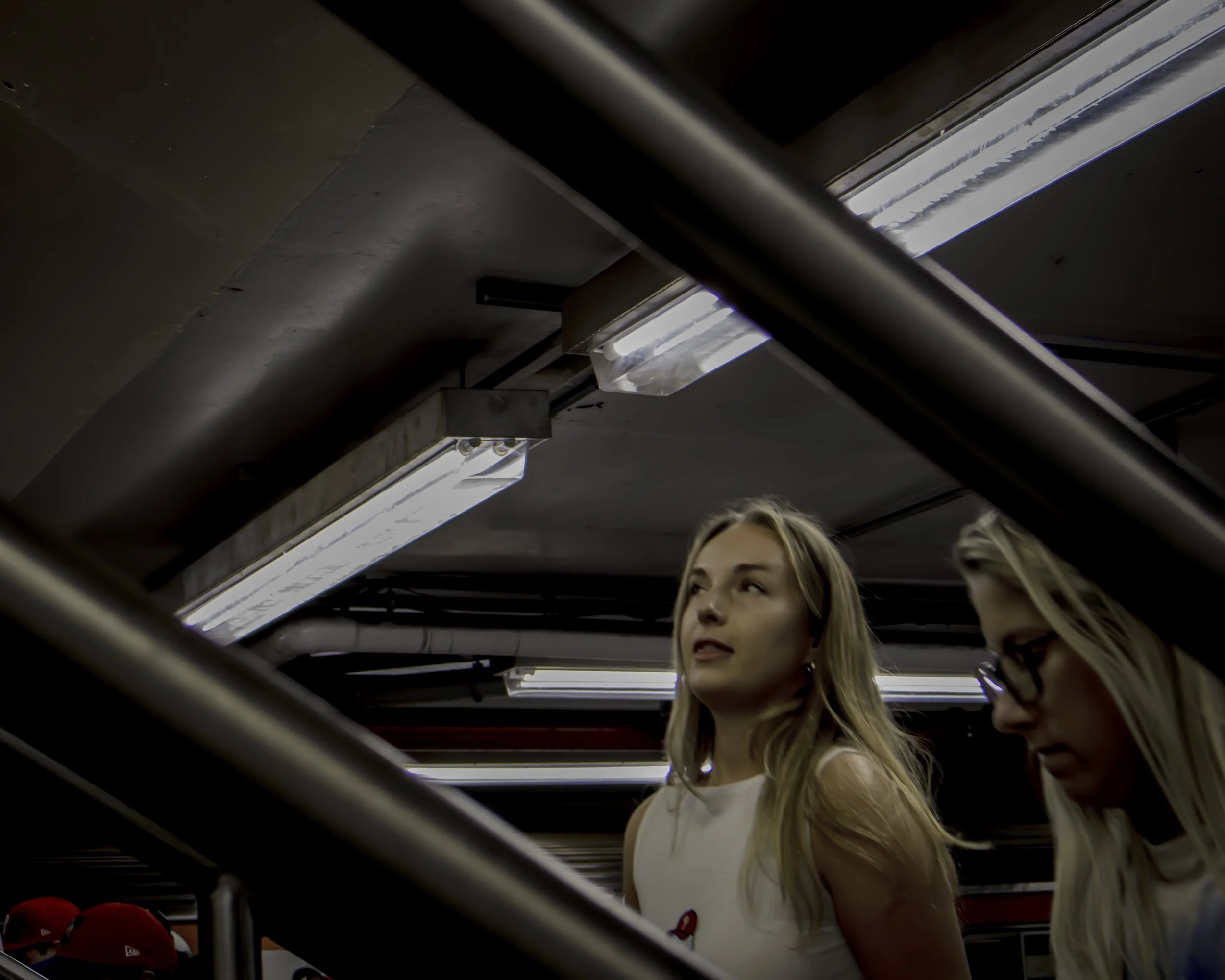 Playoff Anticipation
Two women move through a transit concourse beneath fluorescent lights, framed by diagonal black railings and dark ceiling beams.