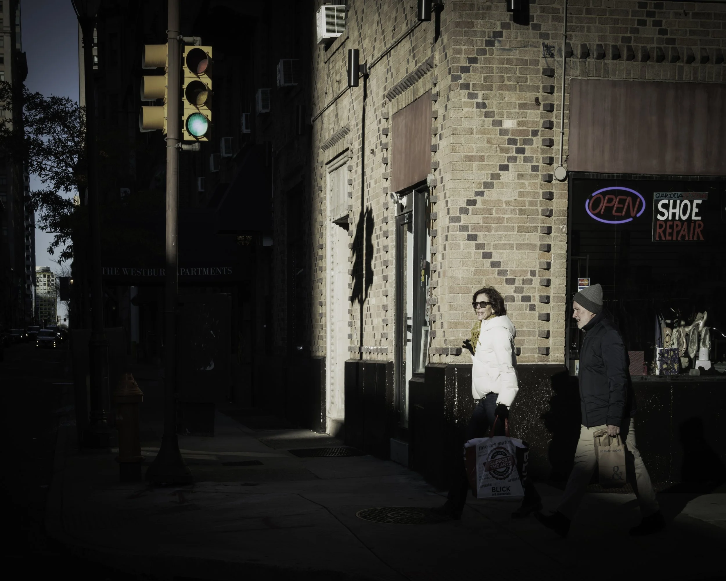 Crossing the Bright Patch
Two pedestrians carrying shopping bags walk through a bright patch of light past a shoe repair shop.