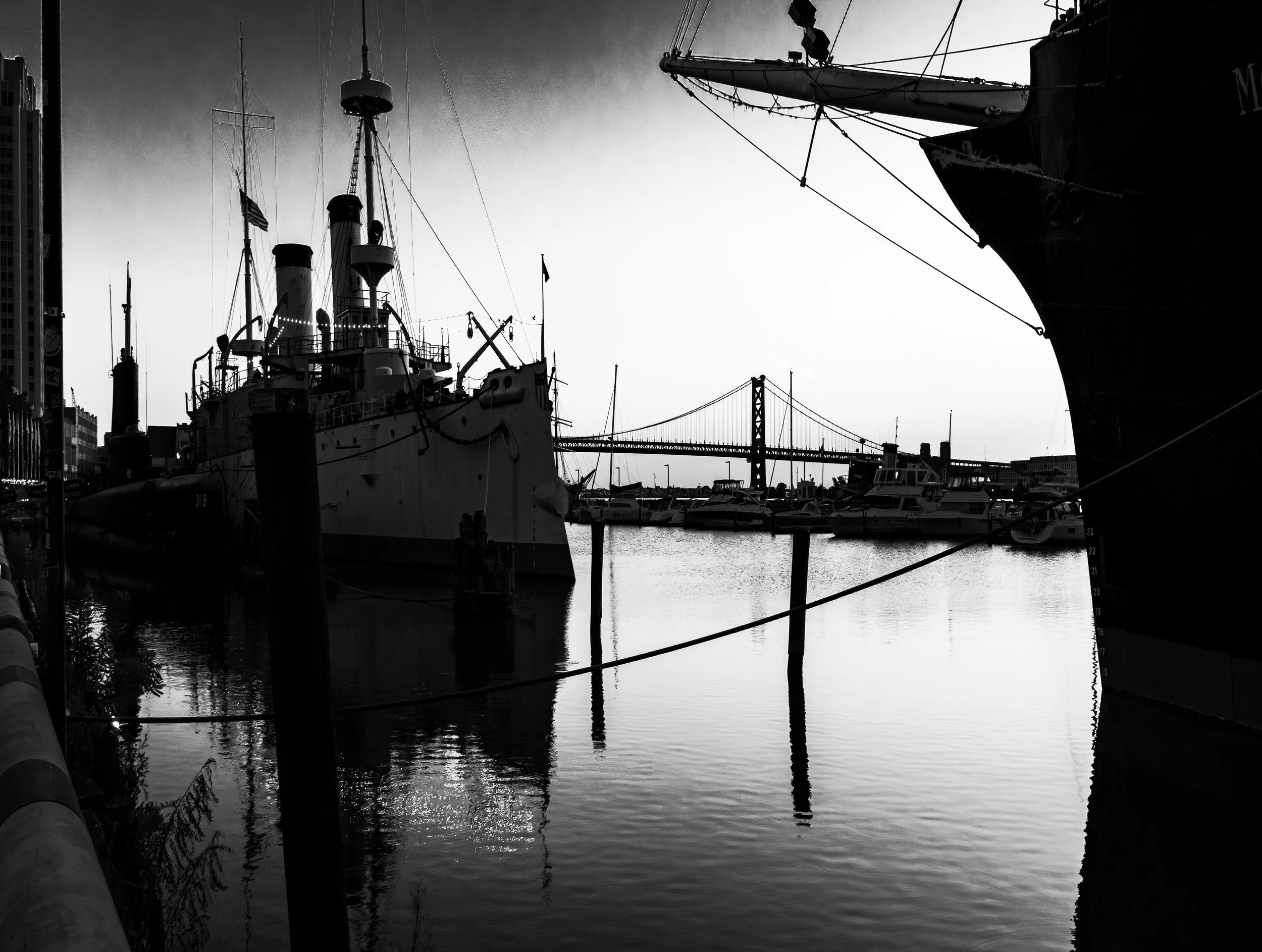 Black and white harbor photograph featuring historic ship with masts on left, suspension bridge in background, calm water with reflections, modern buildings and marina boats, urban waterfront scene