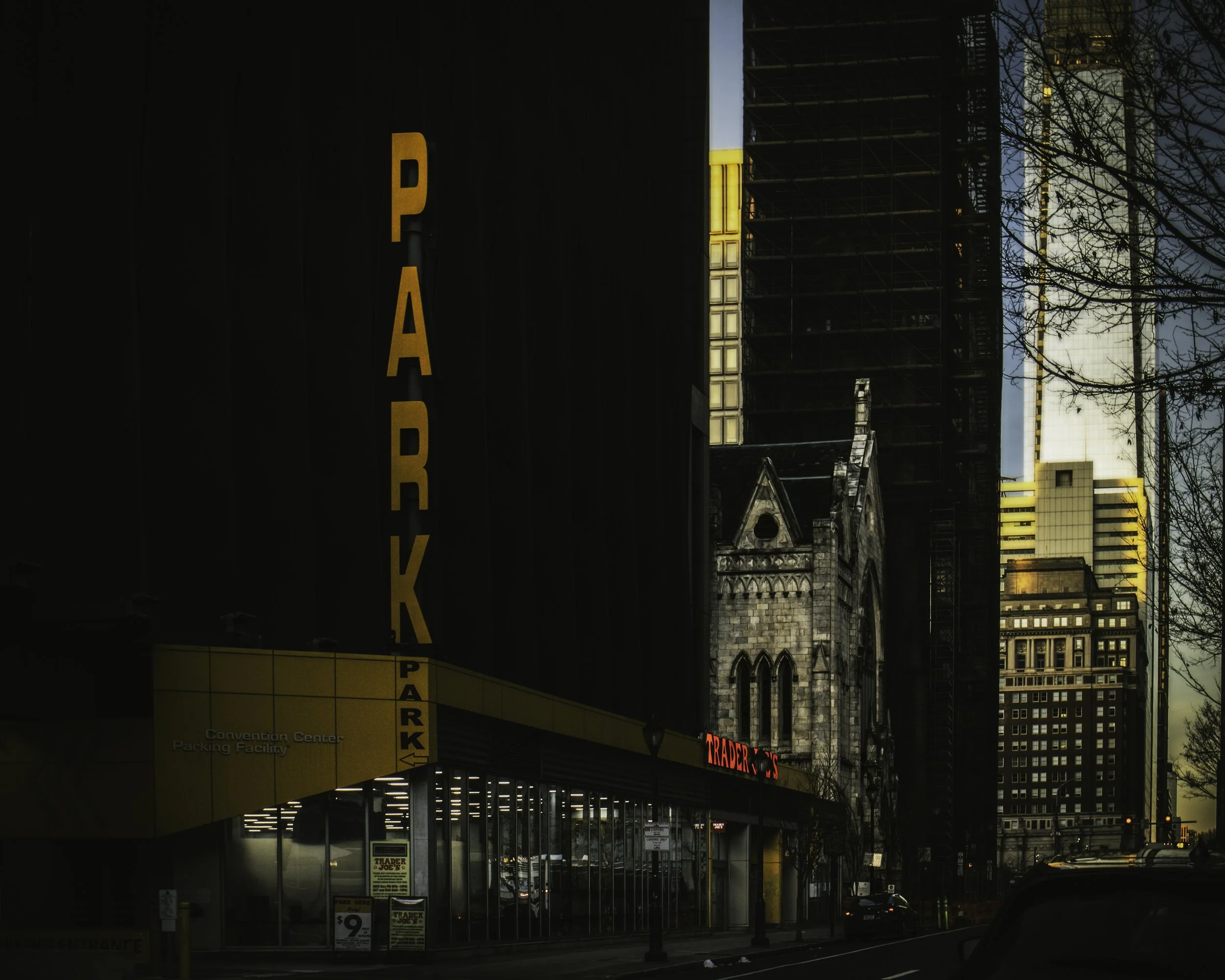 Cinematic downtown Philadelphia street view with a glowing vertical ‘PARK’ sign on a dark garage facade, a yellow parking entrance and storefront windows at street level, and a stone church framed by tall glass-and-steel buildings catching warm sunse