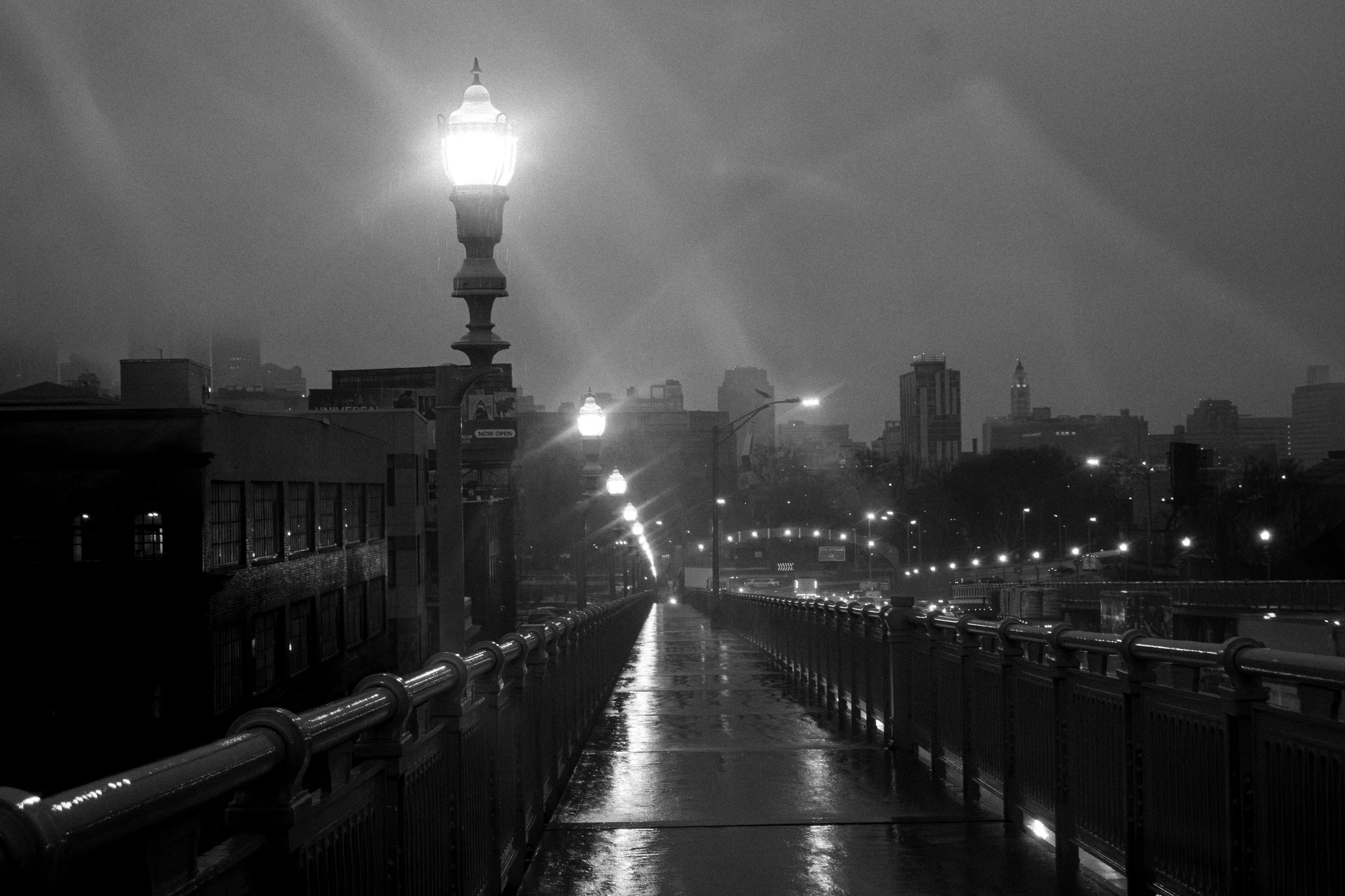 Wet Walk to the City
Black and white photograph of the Benjamin Franklin Bridge walkway in Philadelphia, with glowing streetlamps, wet pavement, and city lights in the distance.