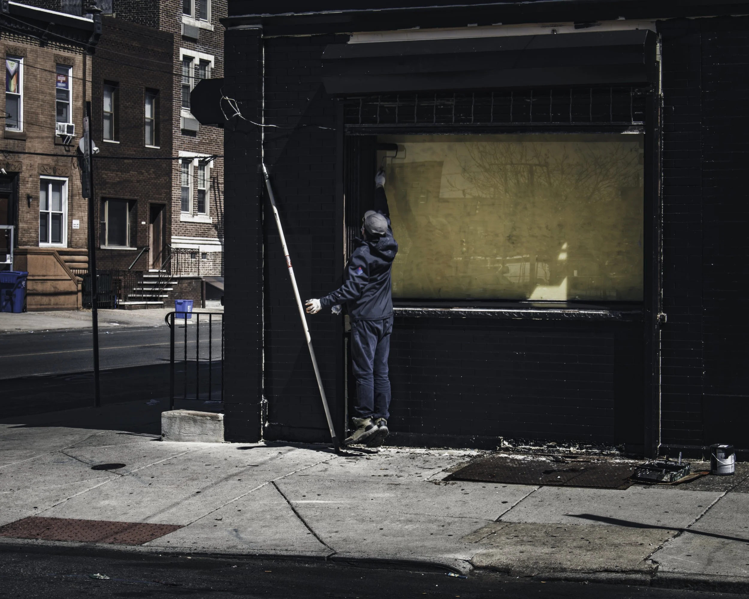 Painting Over Yesterday
A worker in blue coveralls stands on tiptoe against the blacked-out facade of a closed South Philadelphia storefront, reaching up with a long-handled tool while a yellowed boarded window glows behind him, rowhouses and blue re