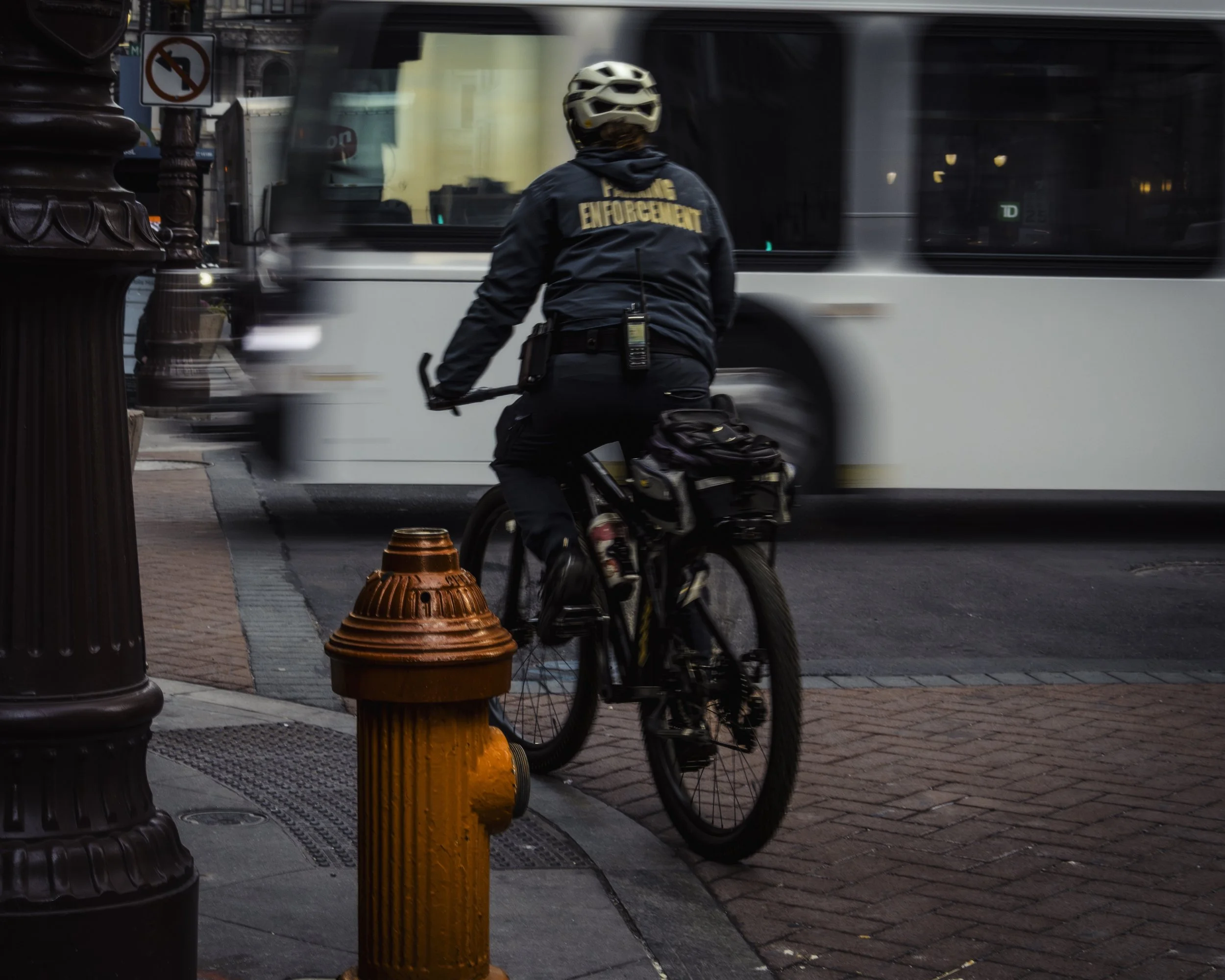 Motion at the Hydrant
Bicycle officer in an enforcement jacket rides beside a blurred bus and an orange fire hydrant at a city corner.