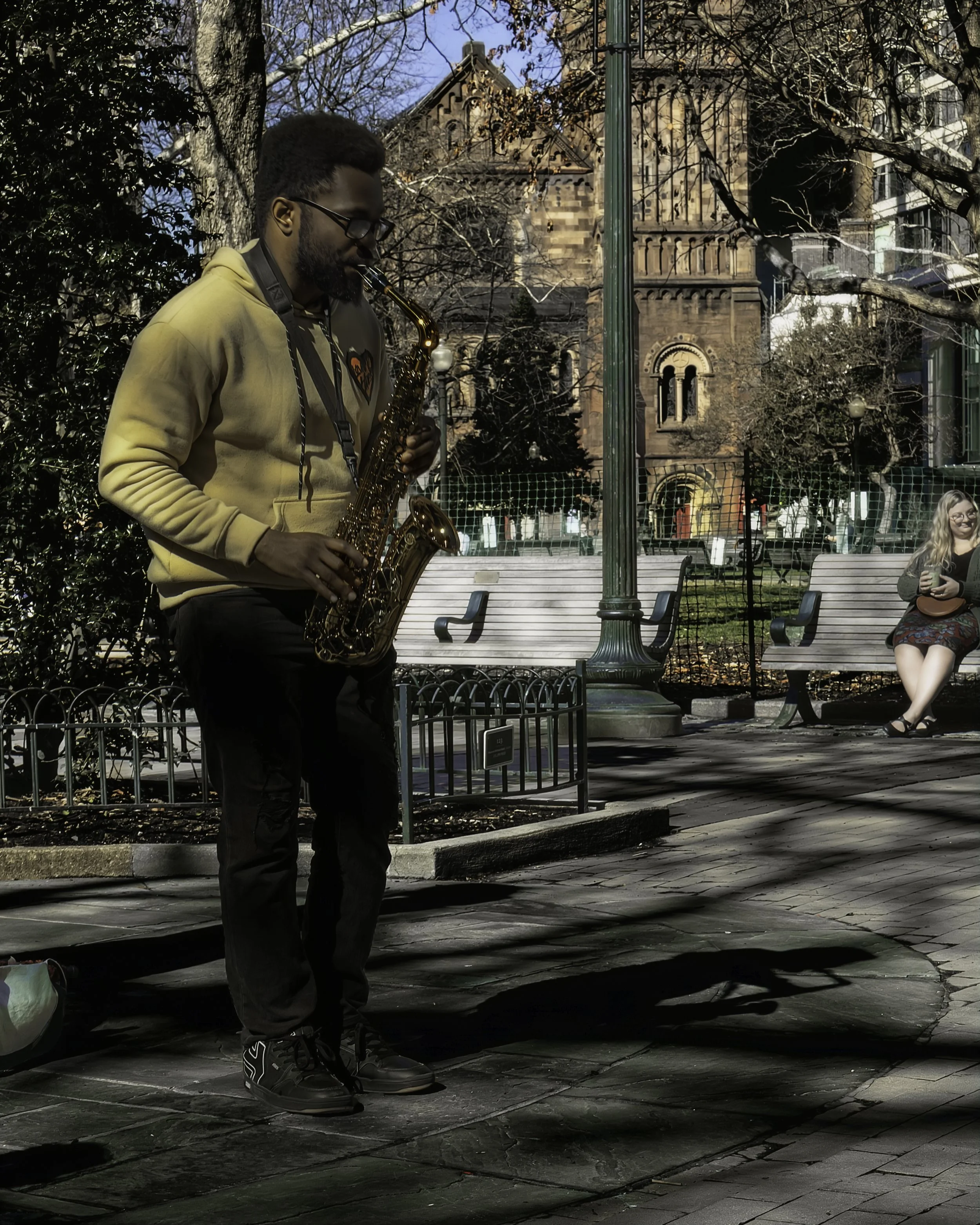 Park Solo
A saxophone player stands in Rittenhouse Park, casting a long shadow of himself playing, while a woman sitting on a bench watches.