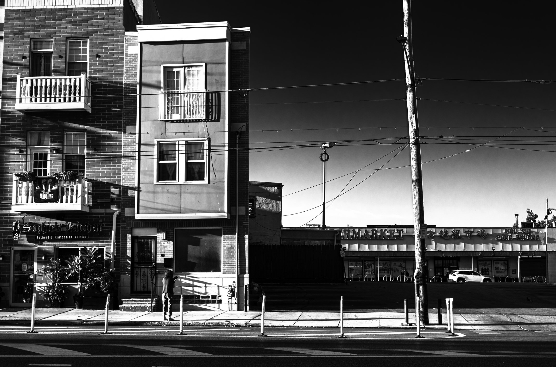 Across from the Market
Black and white street scene along Washington Avenue, with a solitary pedestrian, apartment façades, utility poles, and a market across the road.
