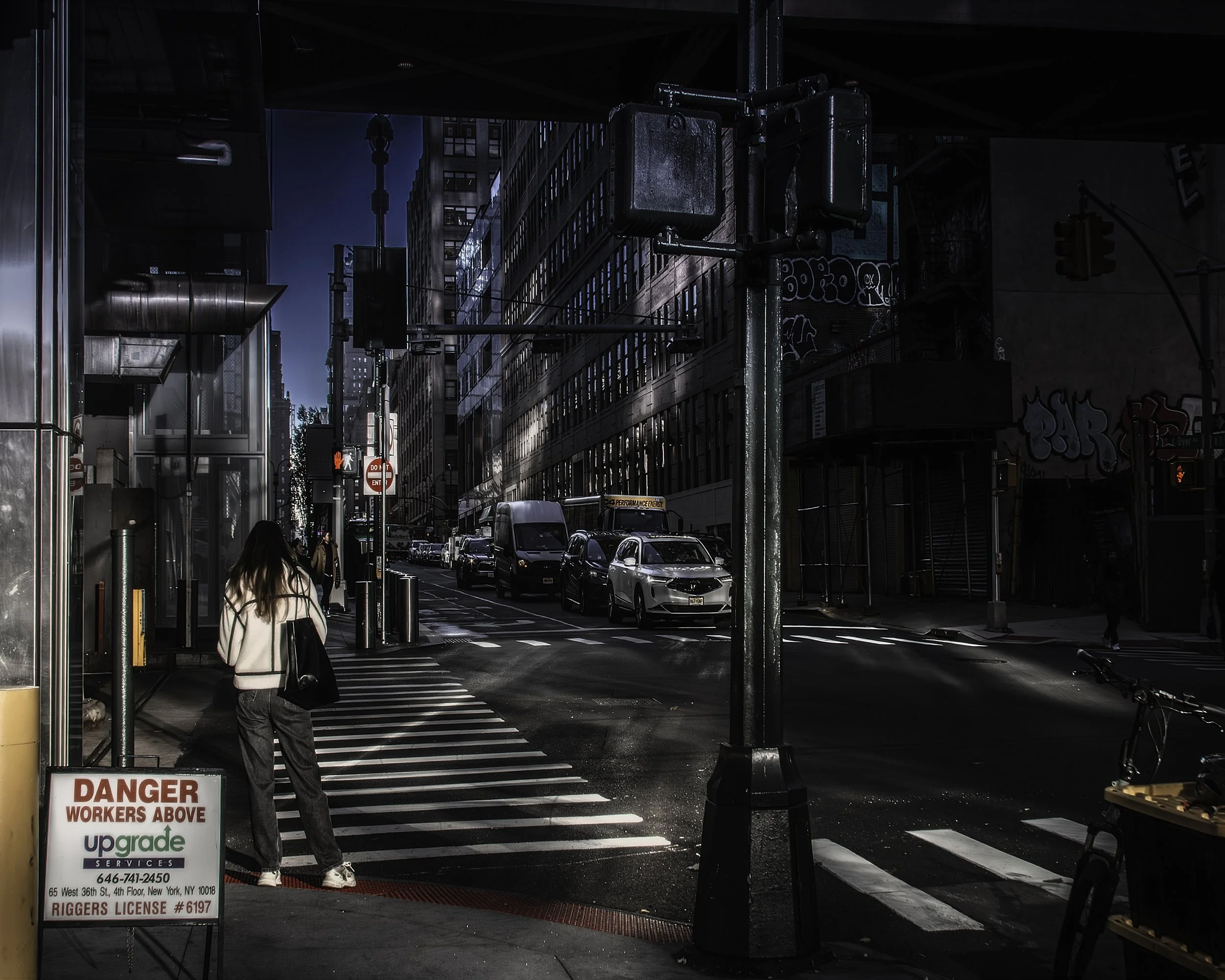 Light Across Thirty-Sixth
Woman stands at a city crosswalk as morning or afternoon light cuts across a wet street lined with tall buildings, traffic, and street signs.
