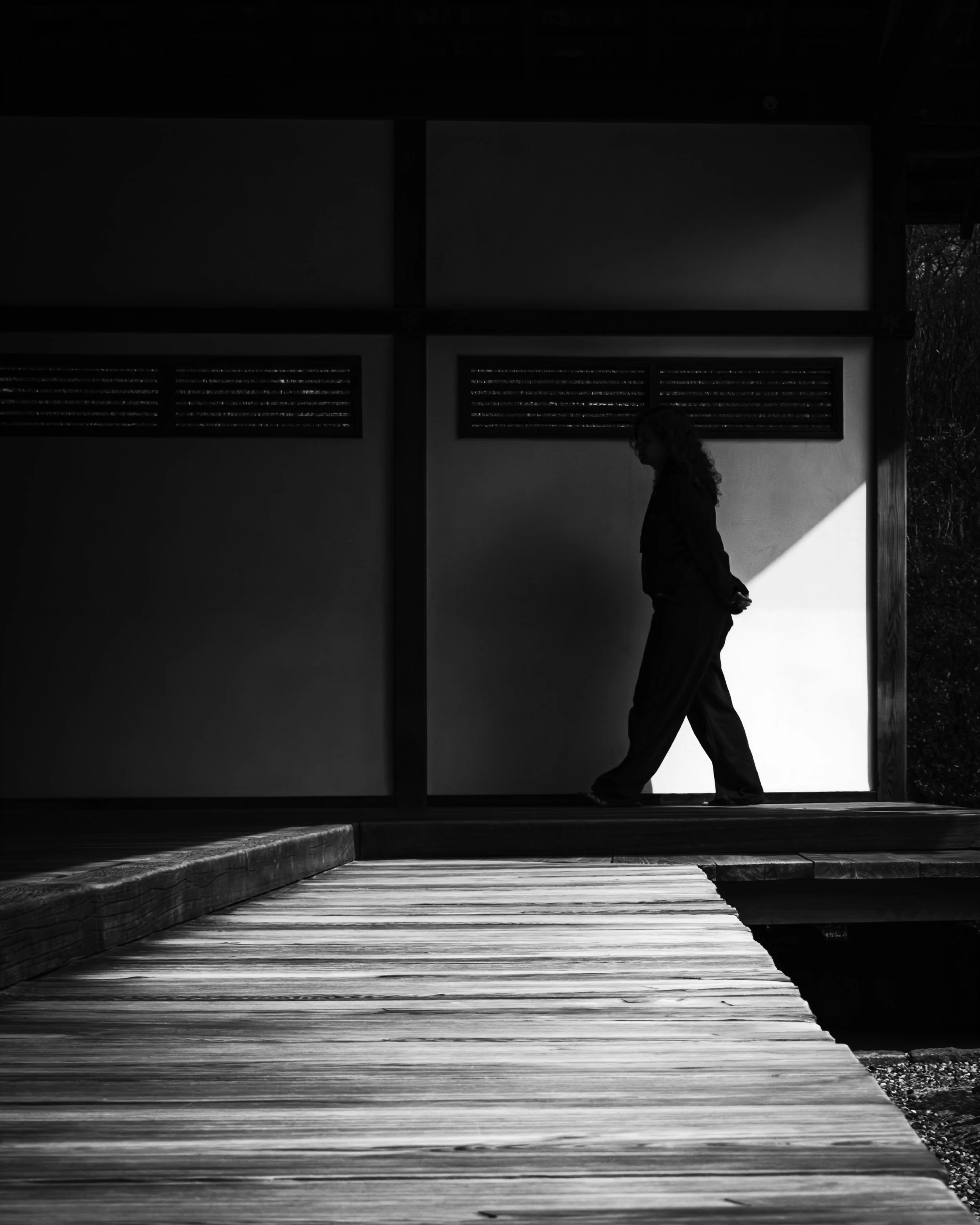 Between Light and Silence
Black and white photograph of a silhouetted person walking at Shofuso Japanese Cultural Center, with a wooden walkway in the foreground and a bright wedge of light behind them.