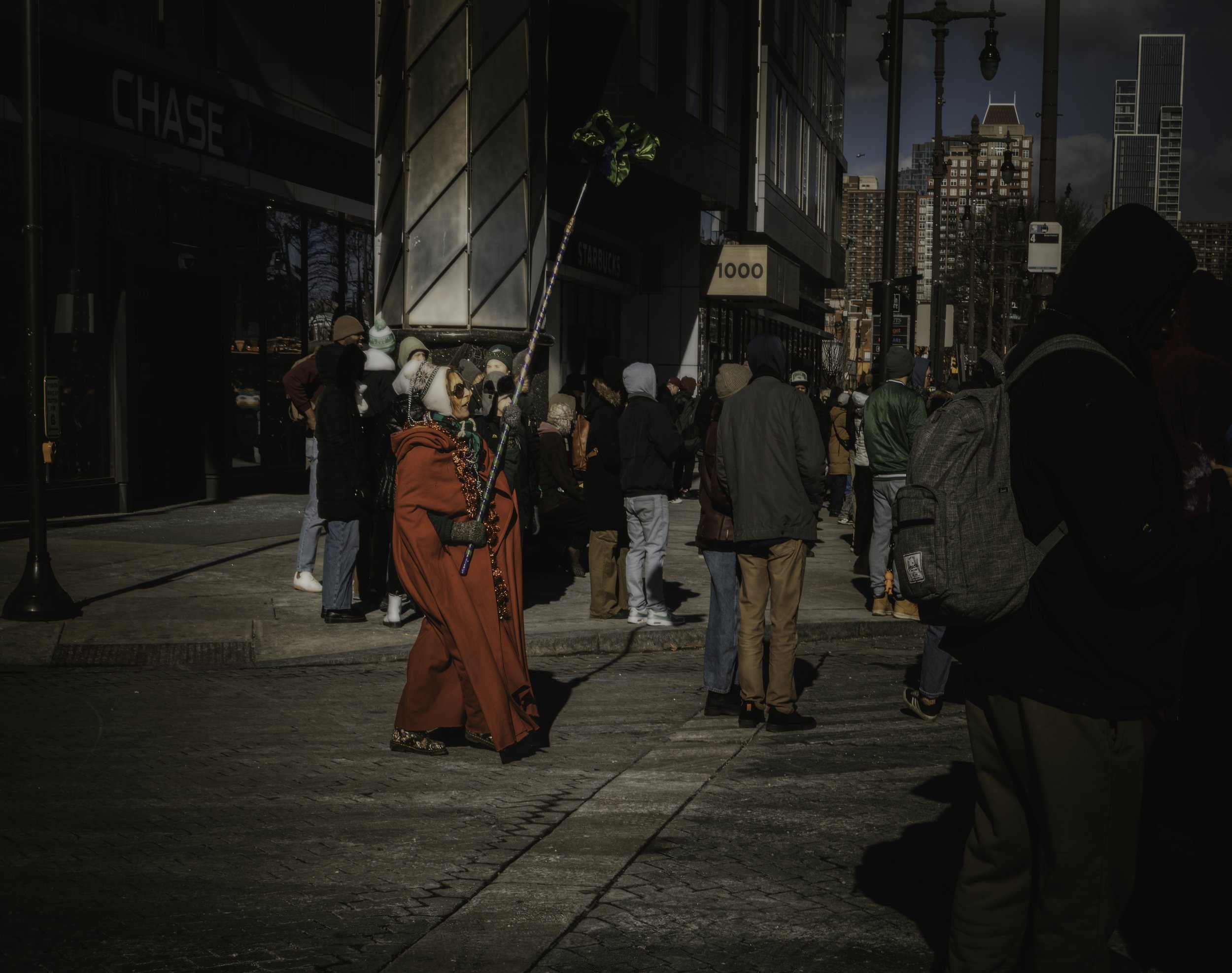 Staff and Spectators
Costumed figure in a red cloak holds a tall staff on New Year’s Day while bundled pedestrians gather at a city intersection.