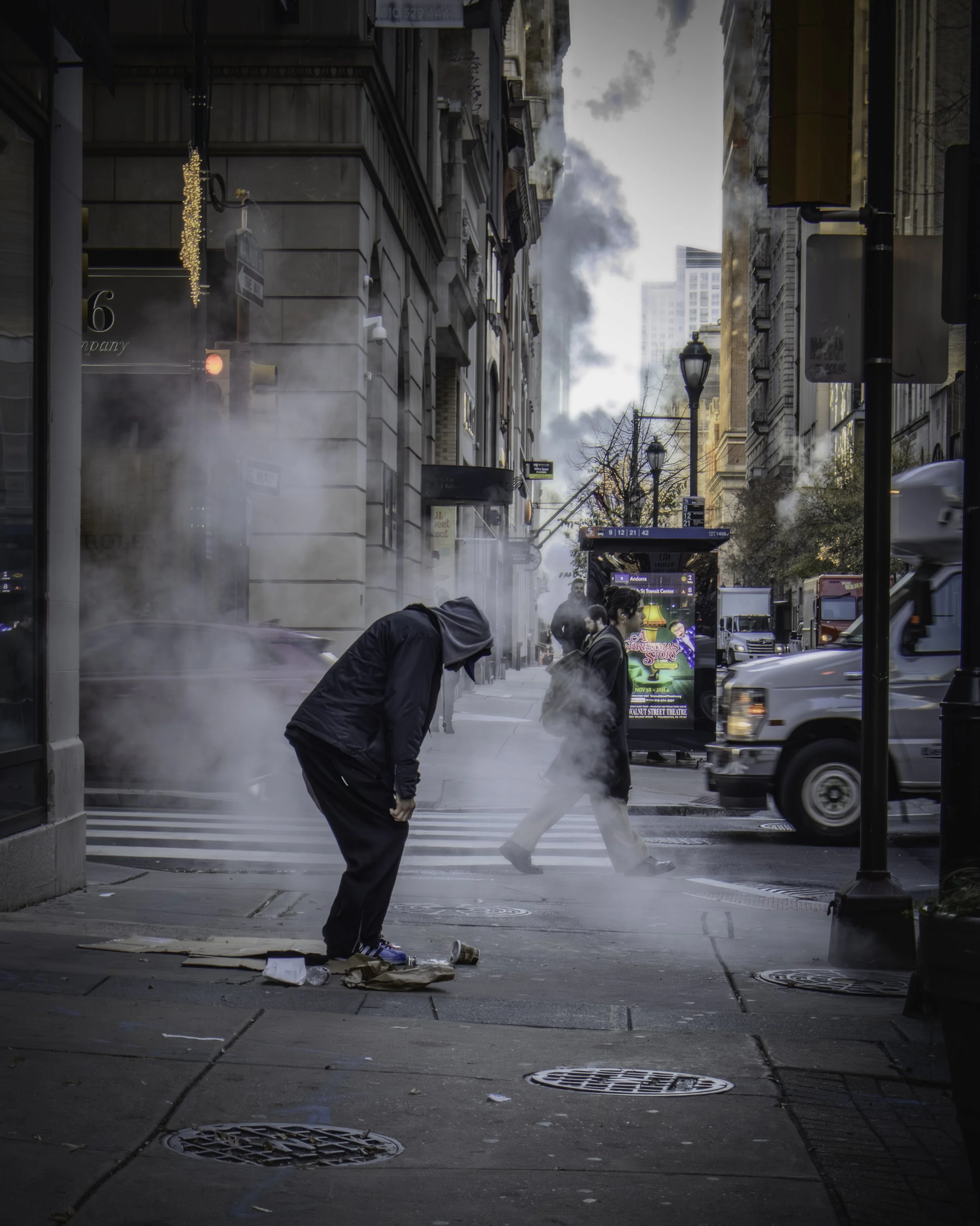 Downtown Exhale
Steam-filled sidewalk scene with a hooded figure bending forward as pedestrians and traffic move through an intersection.