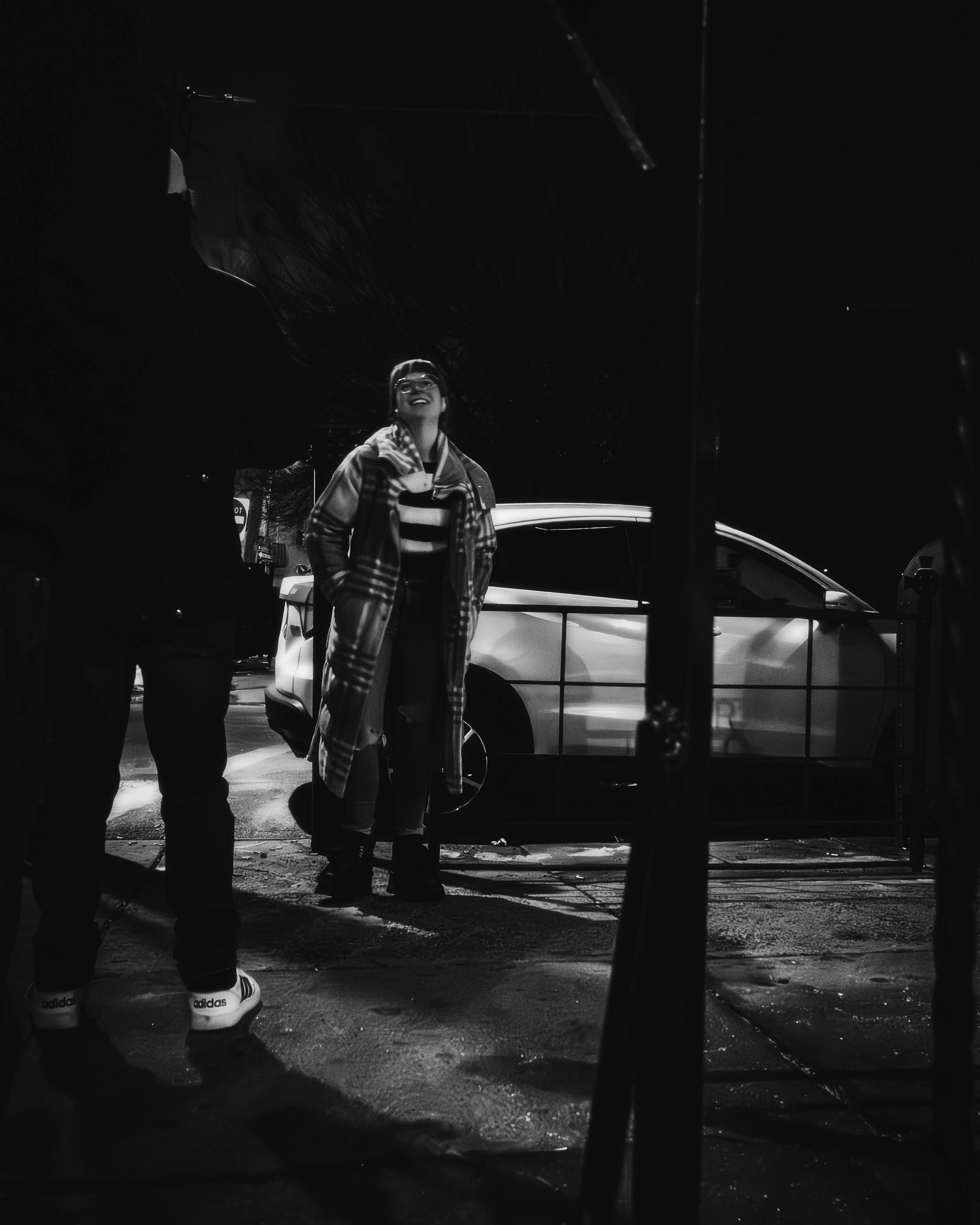 A Bright Moment in Black and White
Black and white street portrait of a smiling woman beside a white car, framed by shadowed figures and wet pavement.