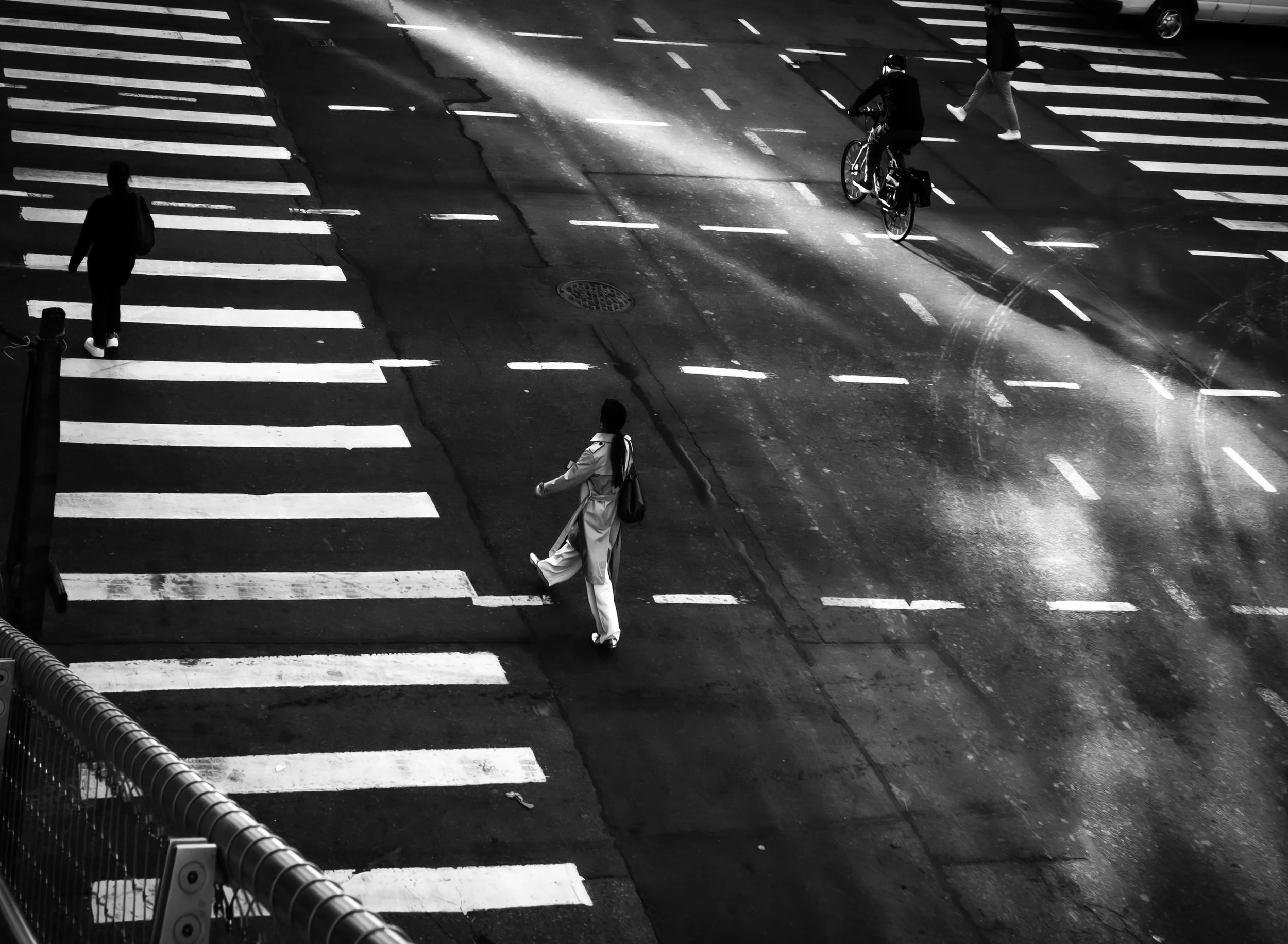 Crossroads in Motion
Black-and-white overhead street scene with bold crosswalk stripes, a woman crossing the road, and a cyclist riding through a bright diagonal band of light.