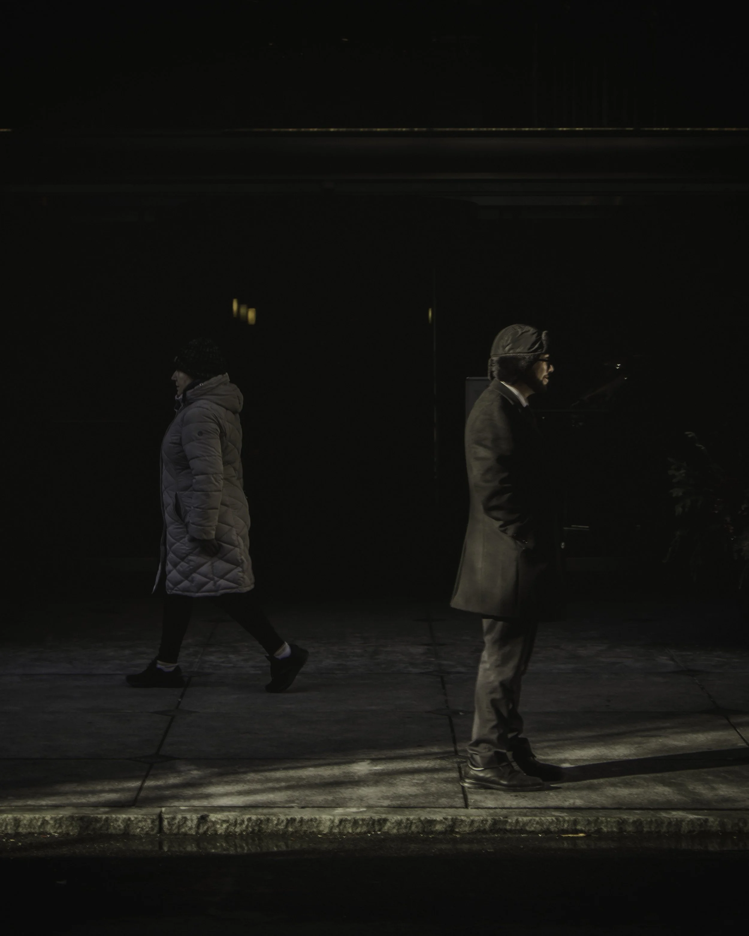 Opposite Directions
Two pedestrians moving in opposite directions on a dimly lit sidewalk, captured in dramatic light and shadow.