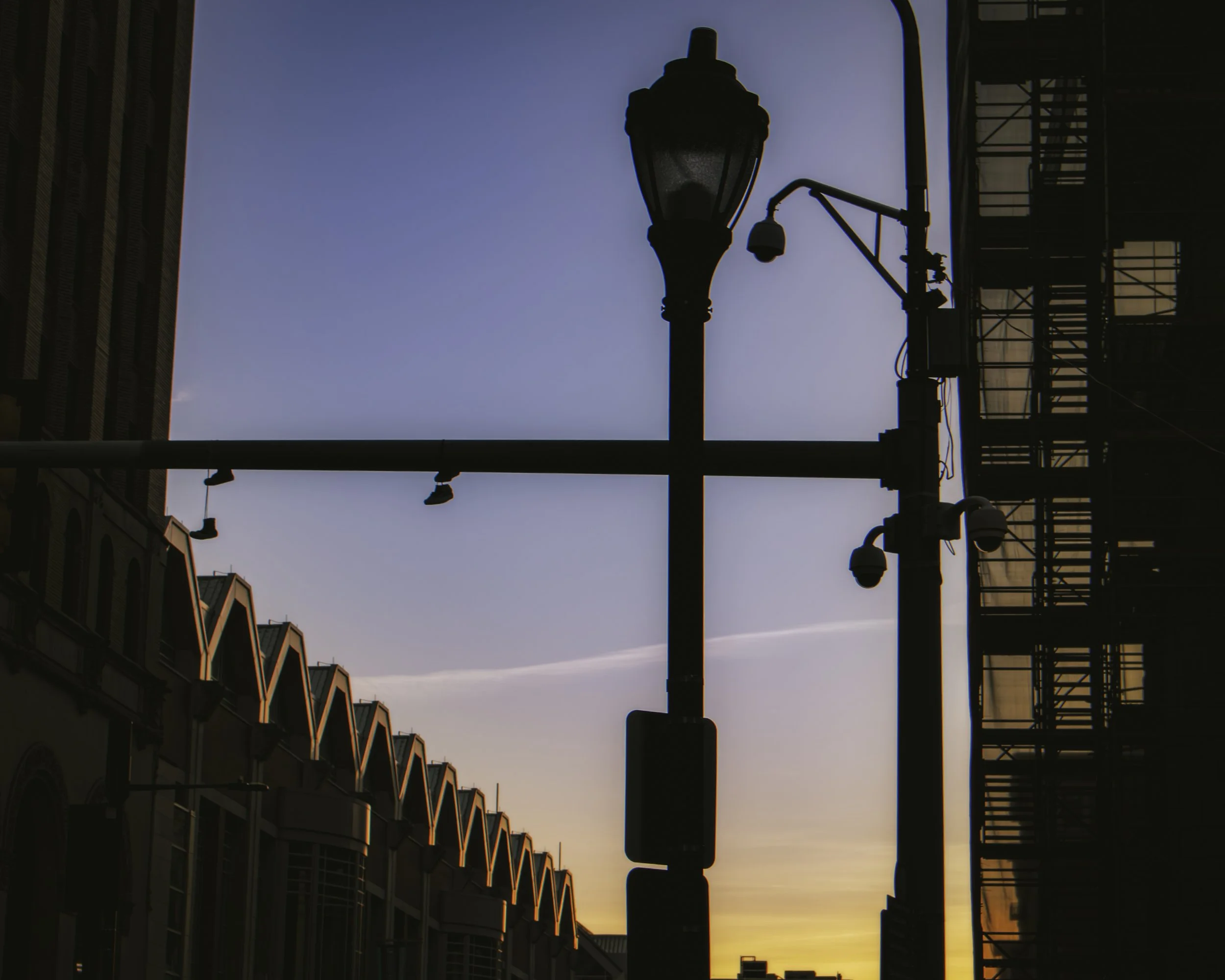 Rooflines at First Light
Silhouetted streetlamp and fire escape beside a row of peaked rooflines at sunrise.
