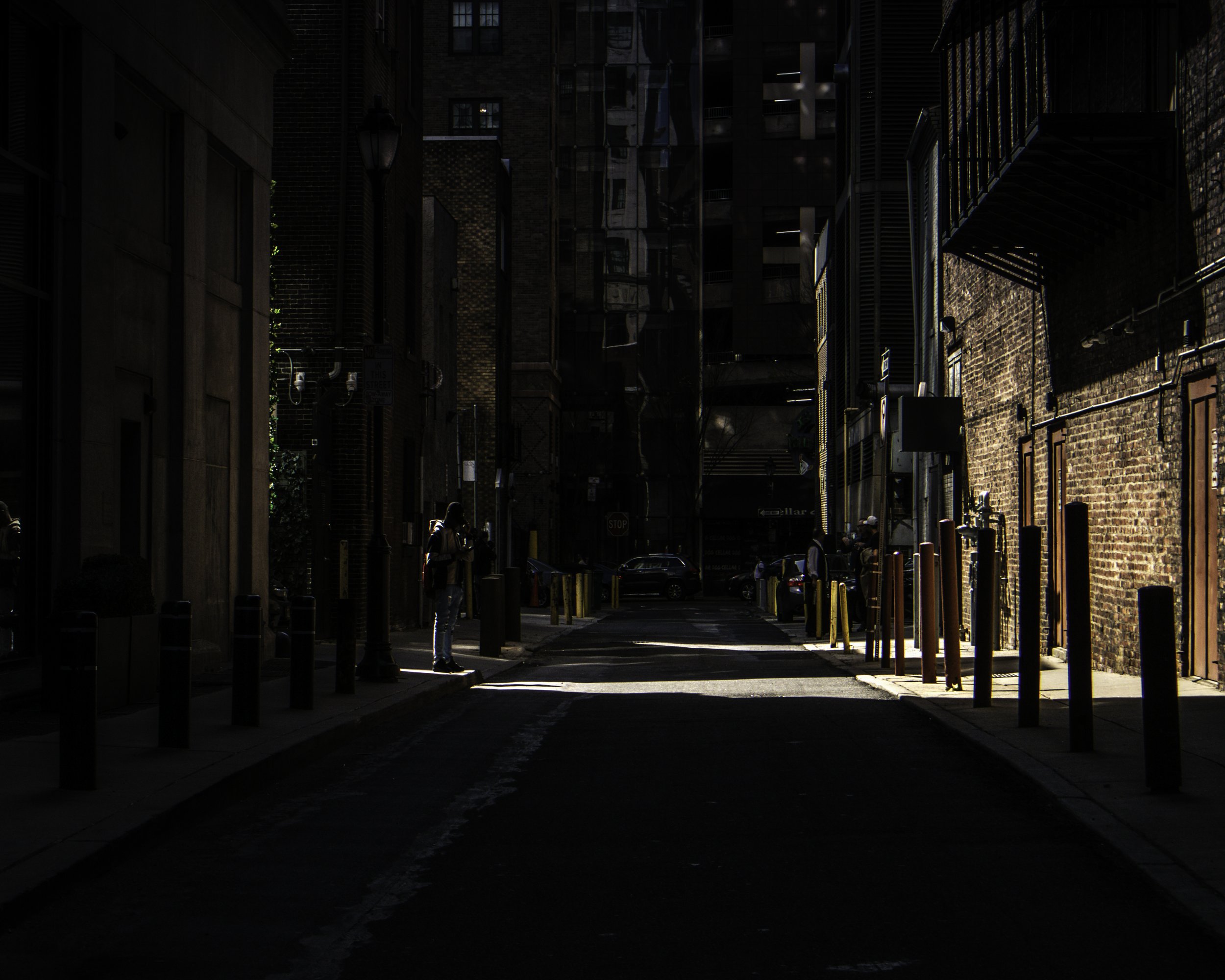 The Only Light on the Block
Near-total darkness fills a narrow Philadelphia alley lined with rows of yellow bollards, a lone hooded figure standing in a single shaft of sunlight mid-left, brick buildings receding into shadow on both sides, a cross vi