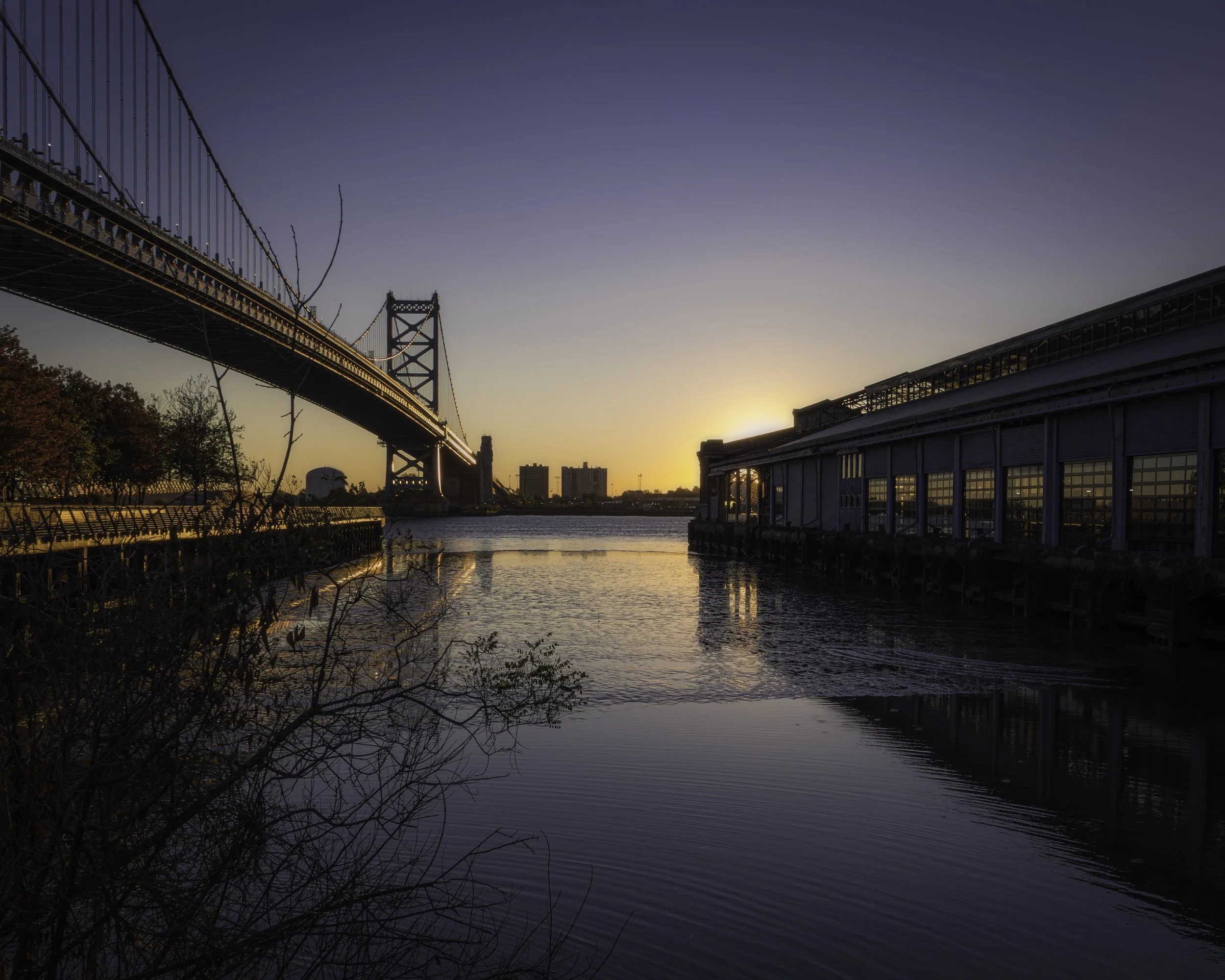 Calm Morning by the Delaware
Sunrise over the Delaware River beside the Benjamin Franklin Bridge, with calm water between the bridge and a long pier building.