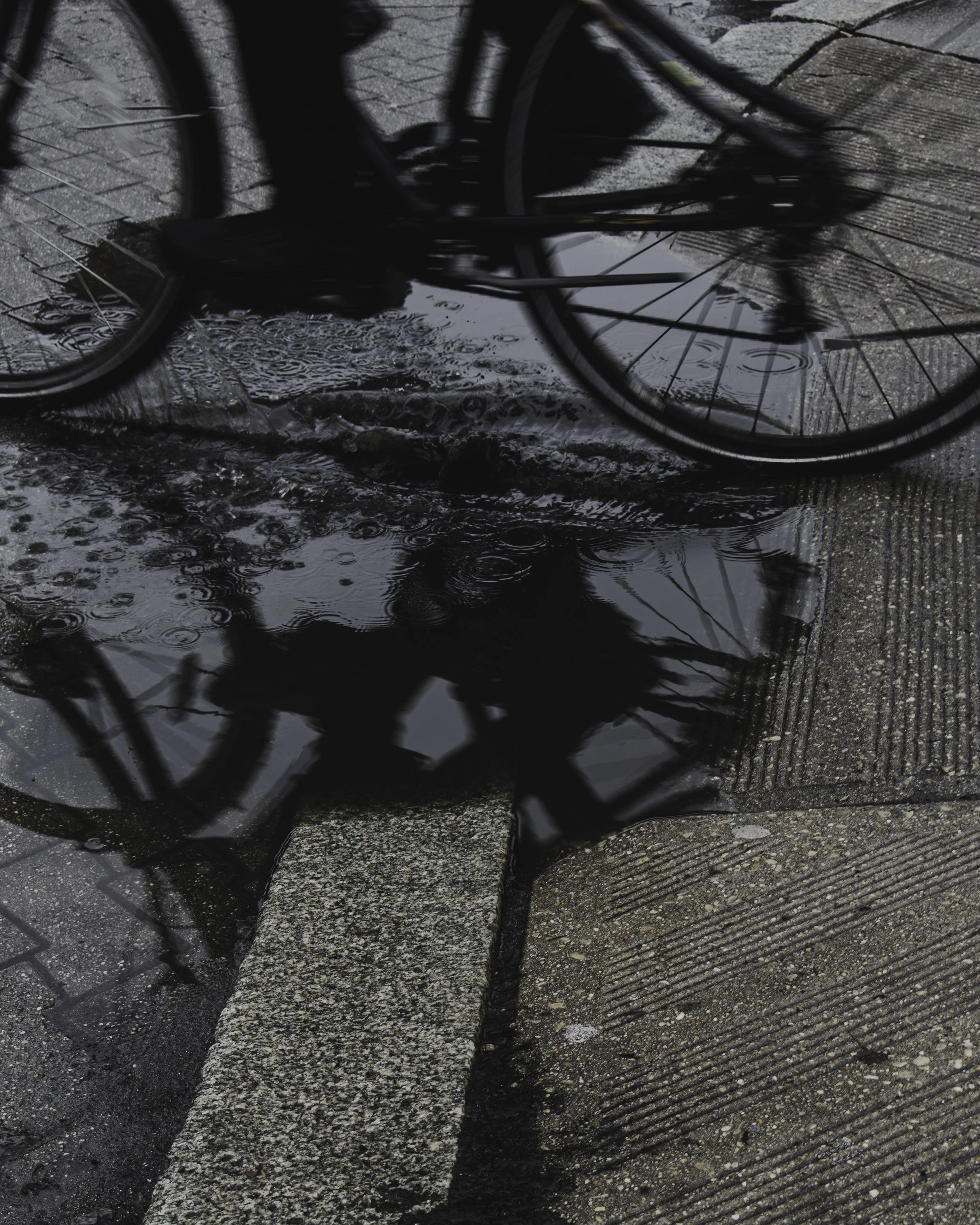 Ripples in Transit
Black and white street photograph of a bicycle passing a rain puddle, with the rider reflected in the water and ripples spreading across the surface.