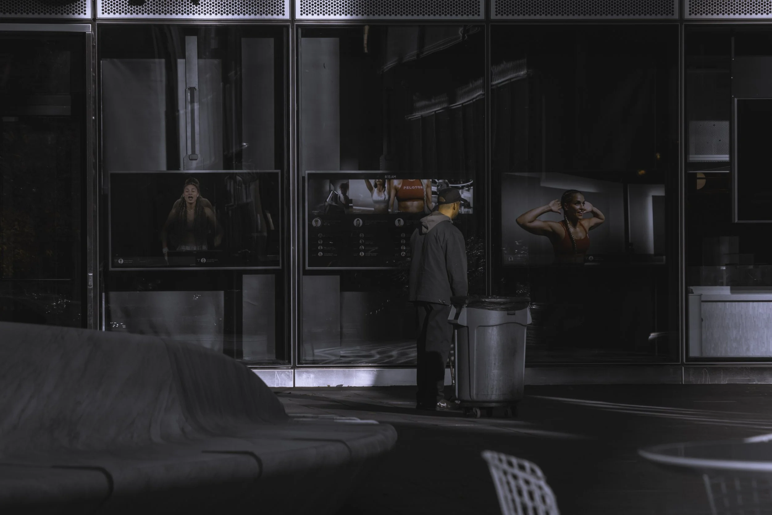 Outside the Glass
Solitary person stands beside a trash bin outside tall glass windows displaying illuminated fitness posters.