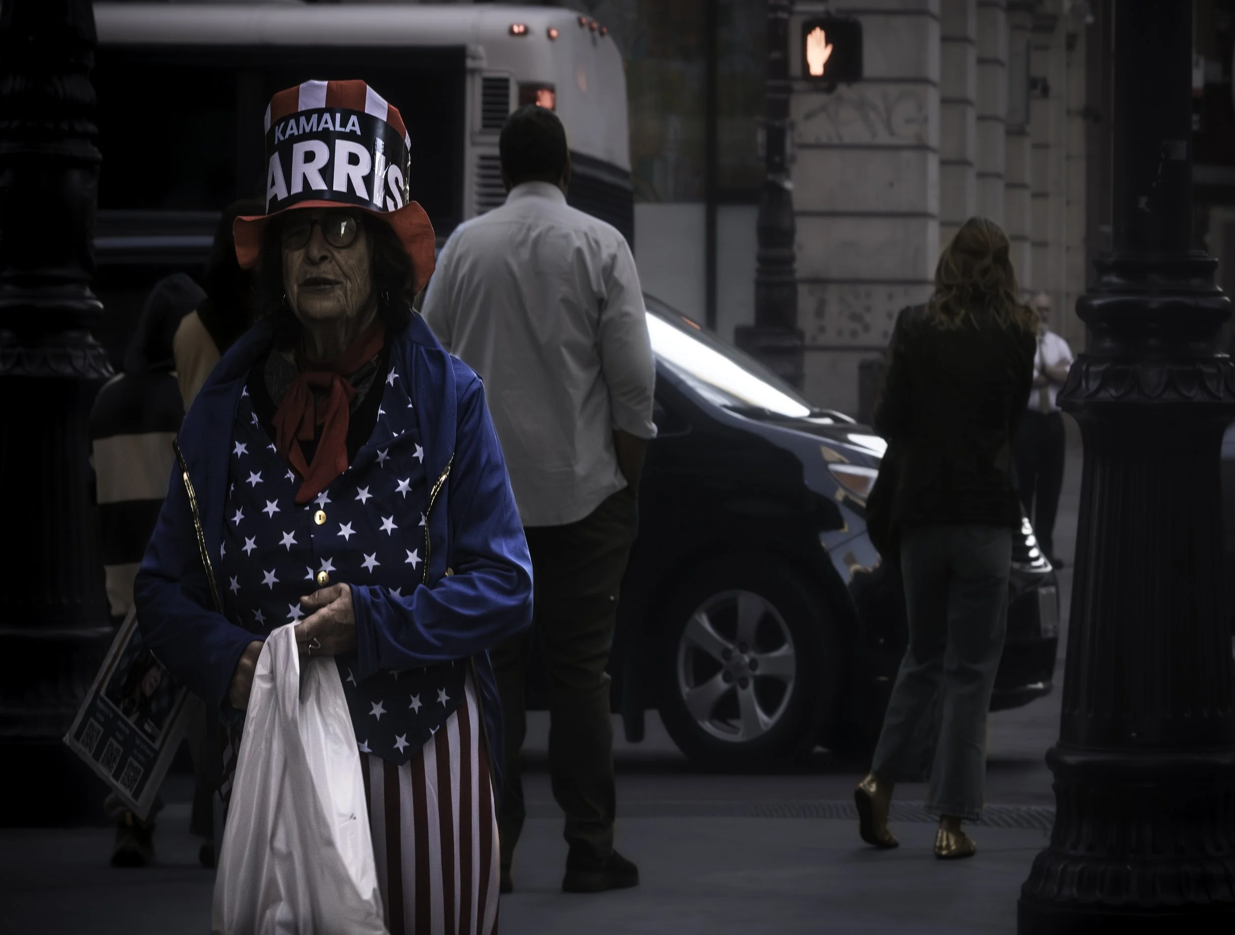 Campaign Hat Figure
Person in a star-spangled outfit and tall campaign hat stands on a city sidewalk holding a white bag.