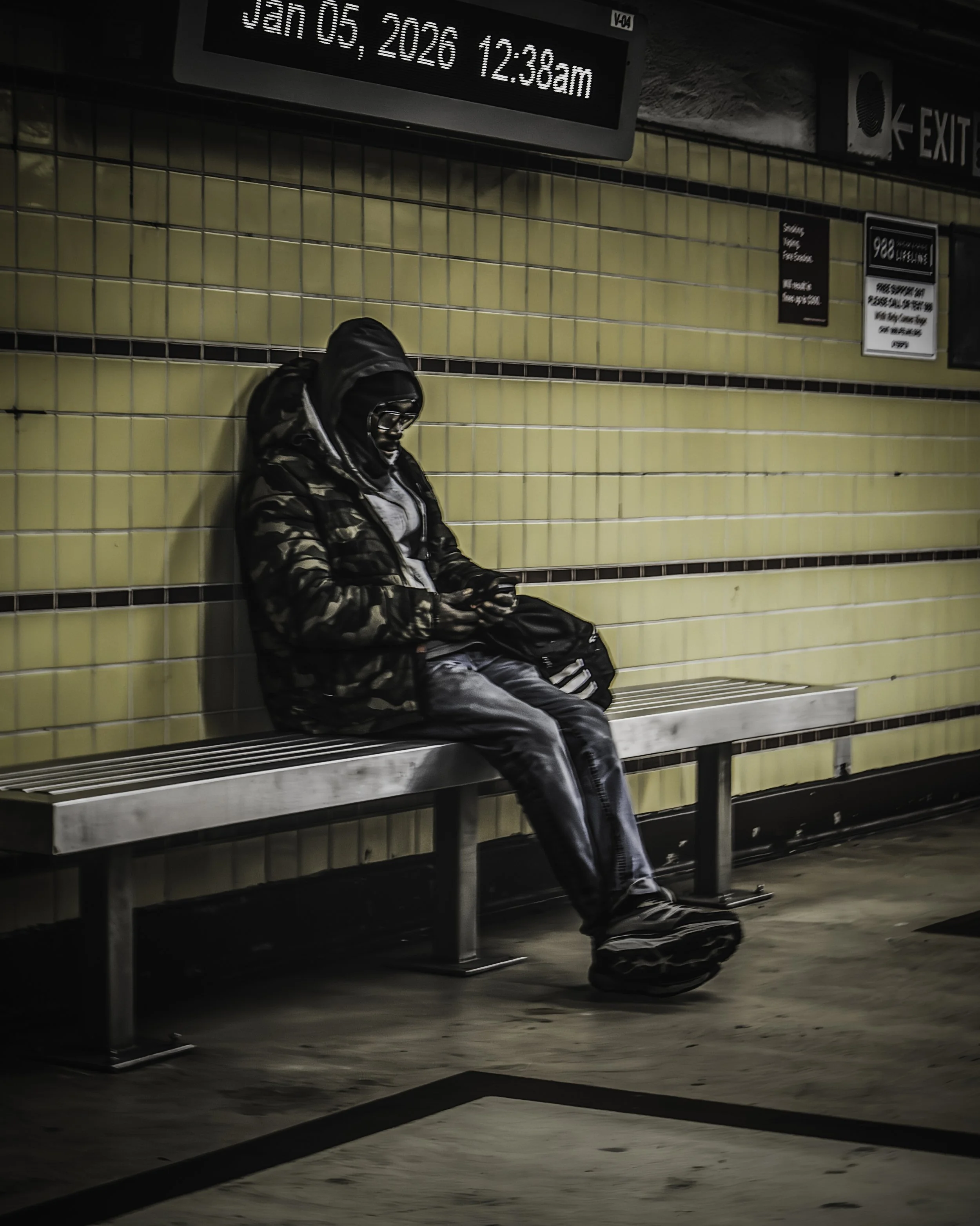 Platform Solitude
Hooded passenger sits alone on a subway platform bench looking down at a phone beneath a digital sign.